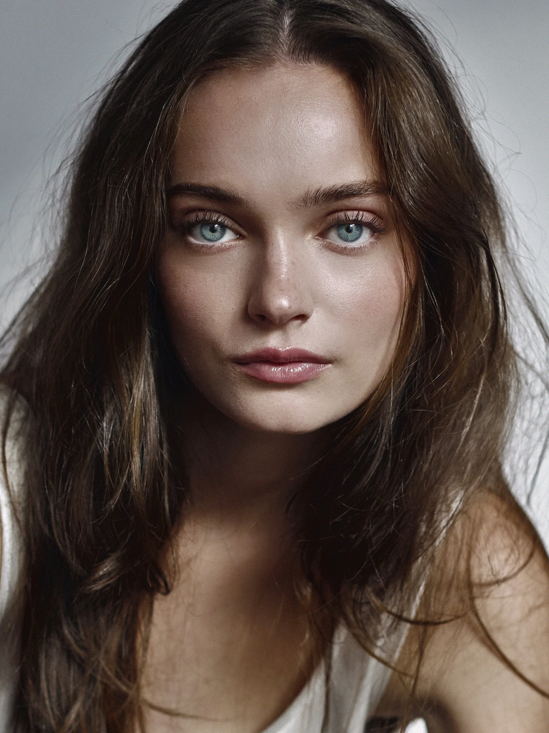 Close-up of a young woman with long wavy brown hair and blue eyes, looking directly at the camera.