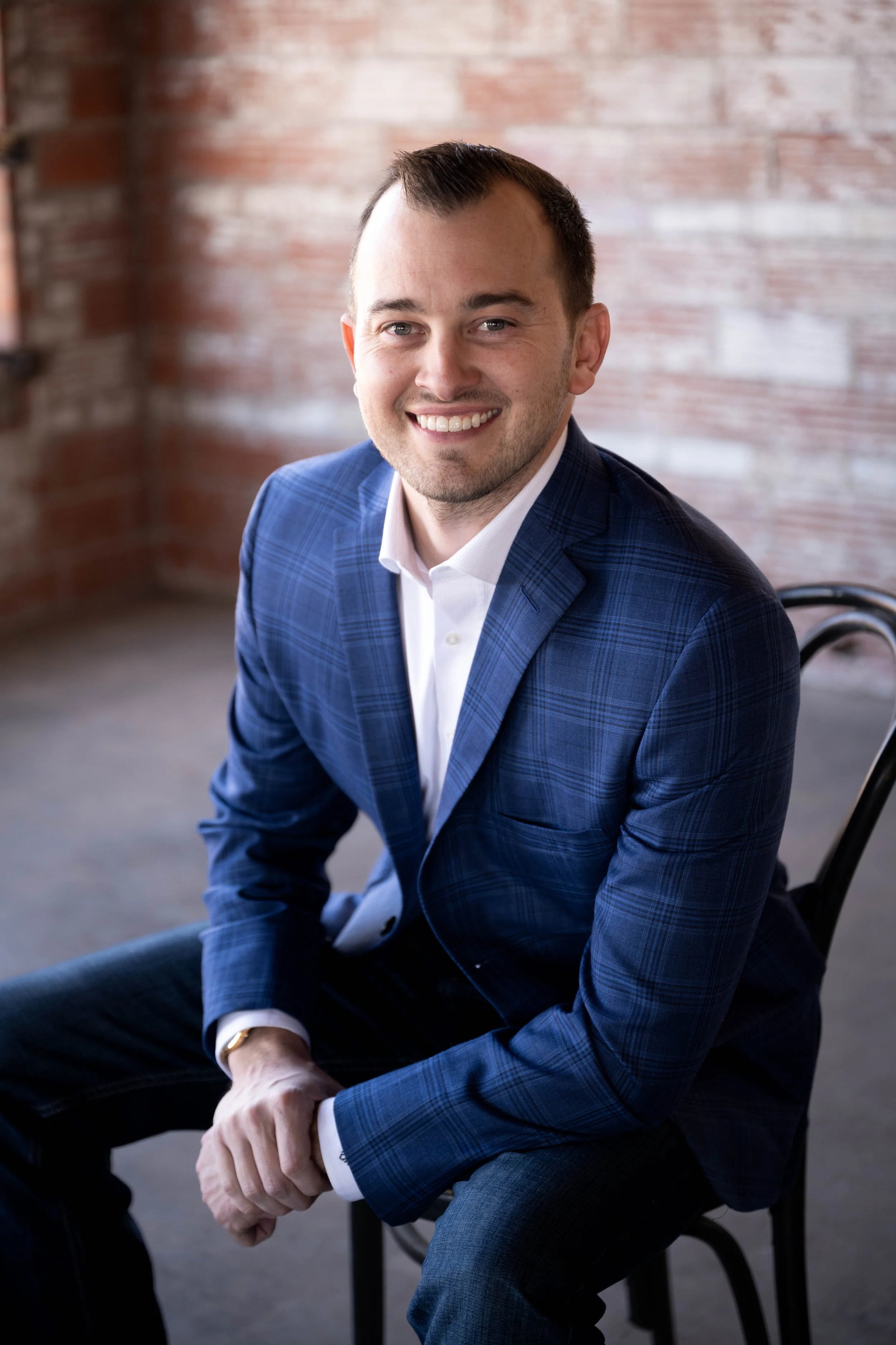 Person smiling, wearing a blue plaid suit jacket and white shirt, seated on a chair against a brick wall.