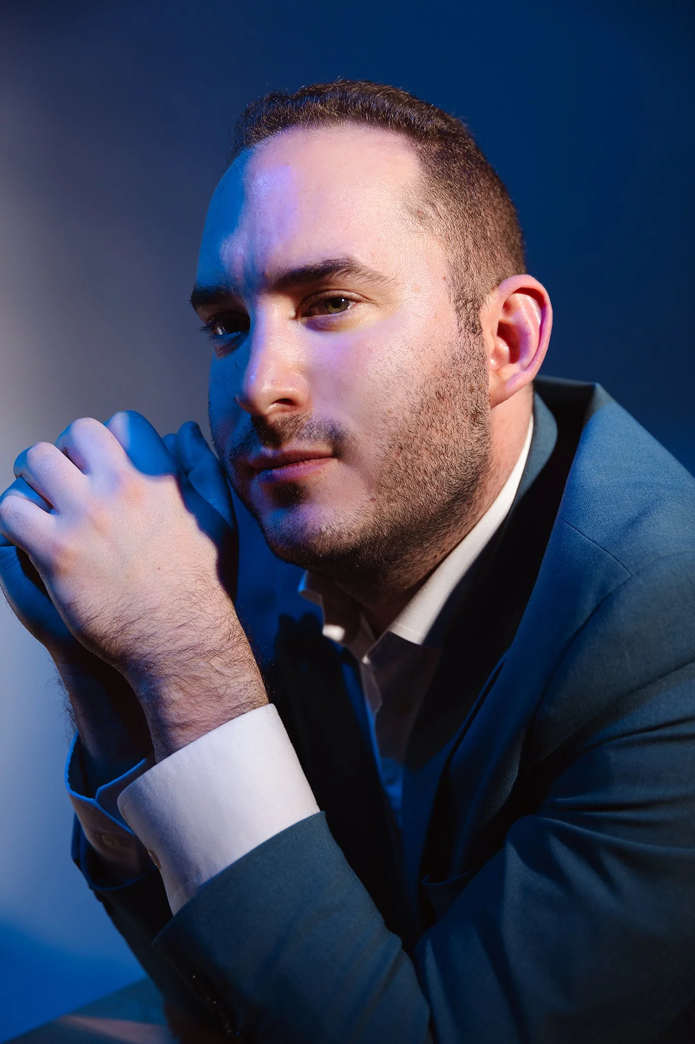 A man in a blue suit with a white shirt, resting his chin on his clasped hands, looking thoughtfully at the camera with a serious expression, against a dark blue background.