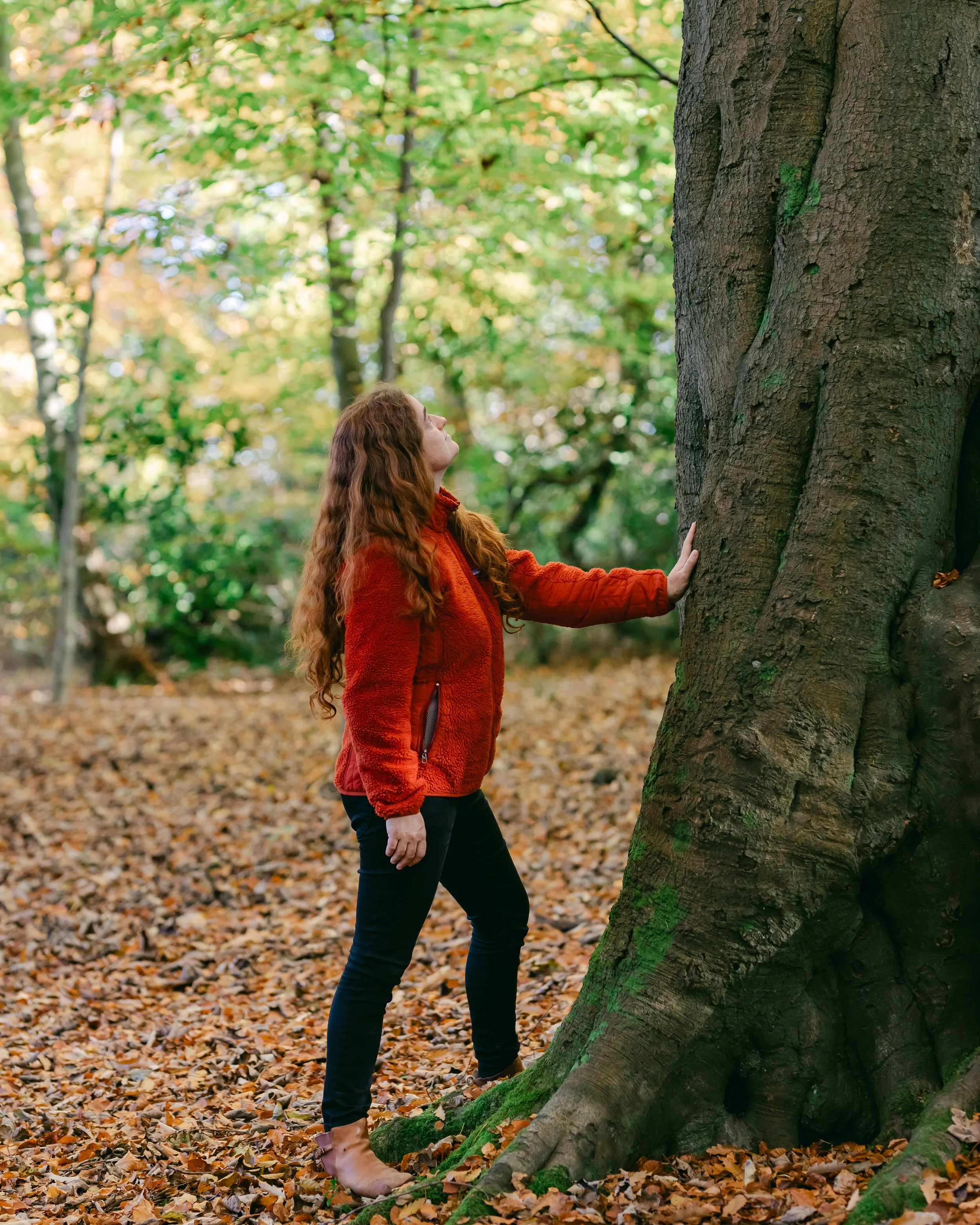 A woman with long red hair in an orange jacket and black pants touches a large tree in an autumn forest.