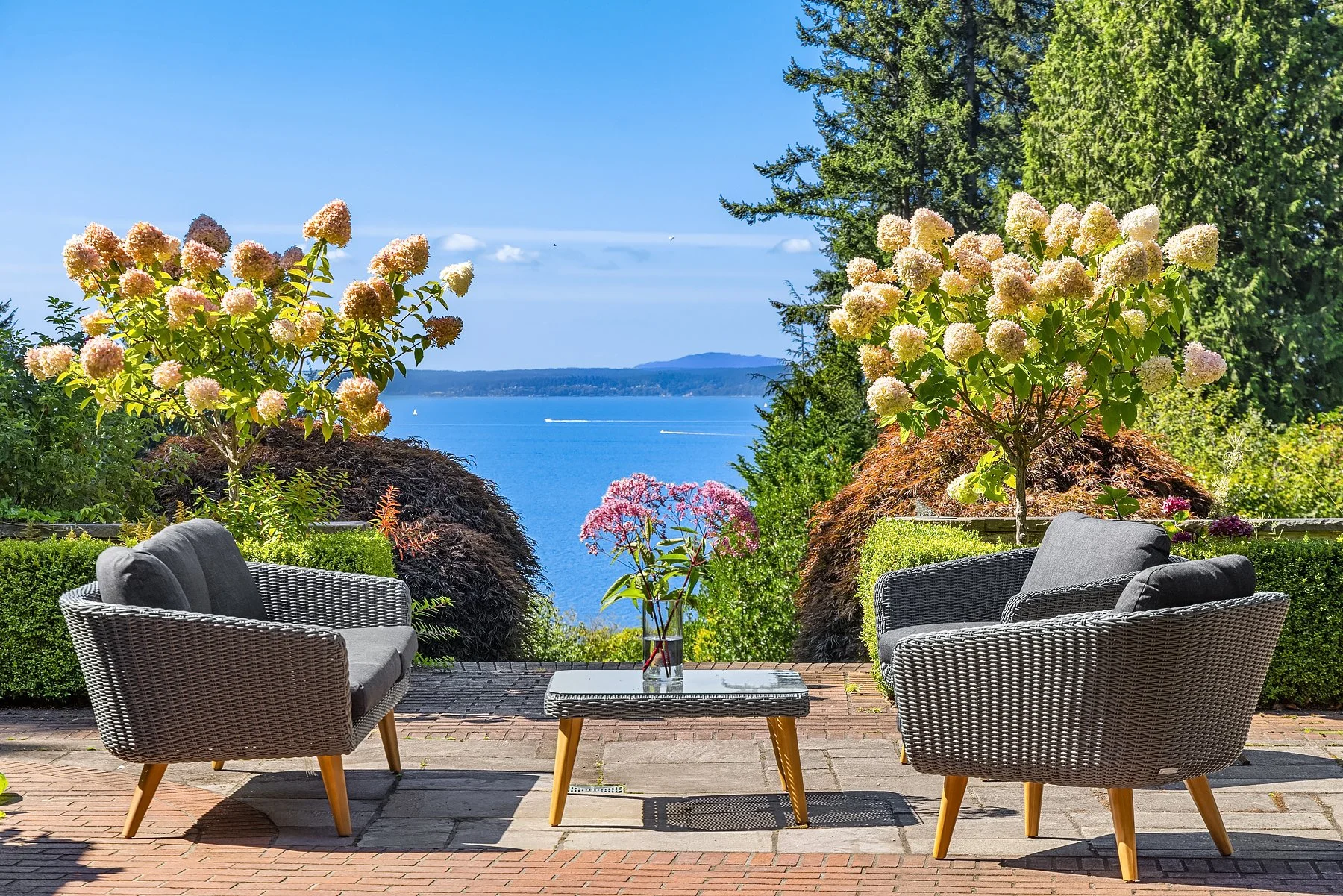 Lush green lawn with trees, a house partially visible, and four empty chairs facing the water, with a view of the ocean at sunset.