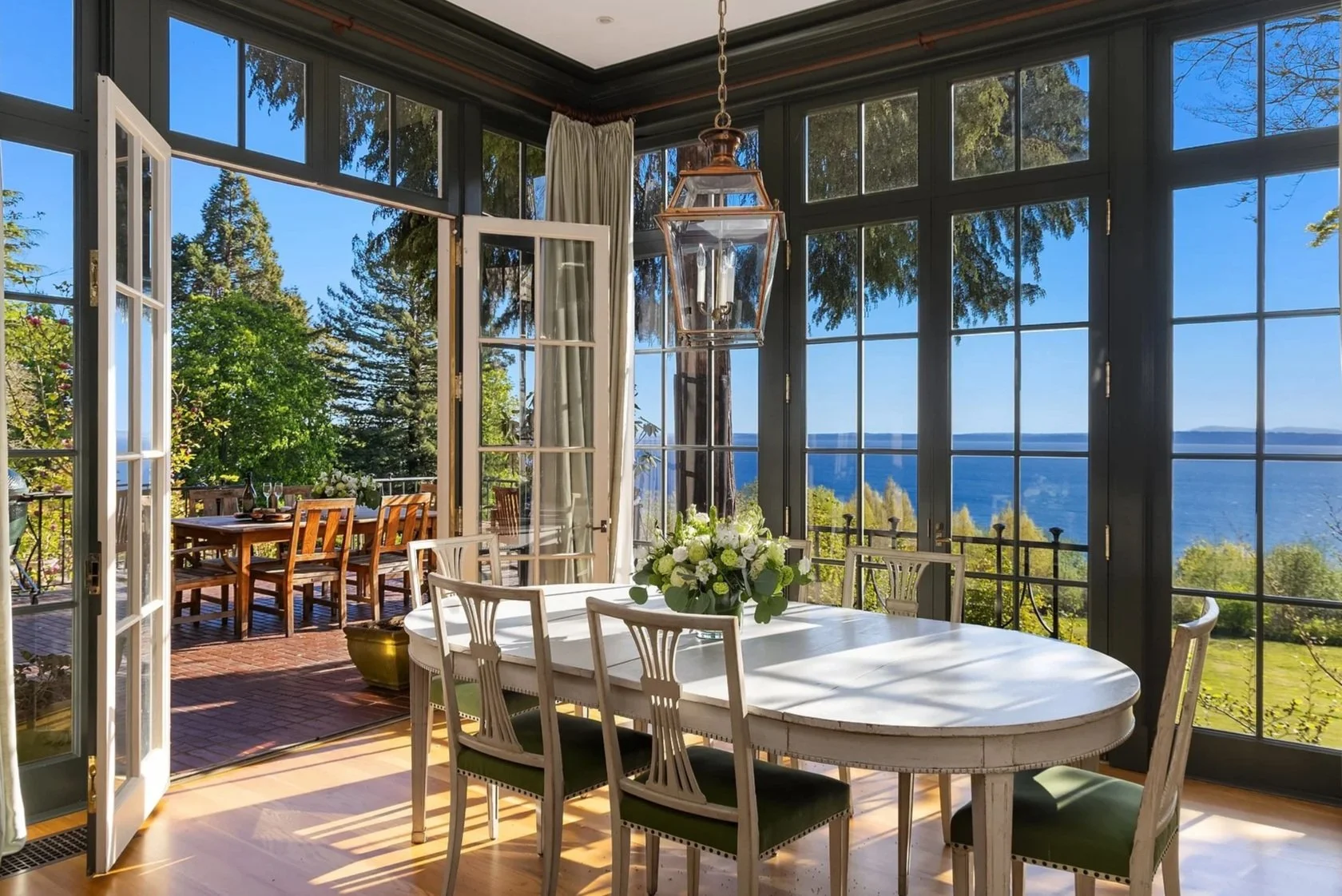 A large, well-maintained house with a tiled roof, surrounded by lush green trees and manicured lawn, with a curved driveway leading up to it. In the background, there are tall forests and a view of a blue body of water under a clear sky.