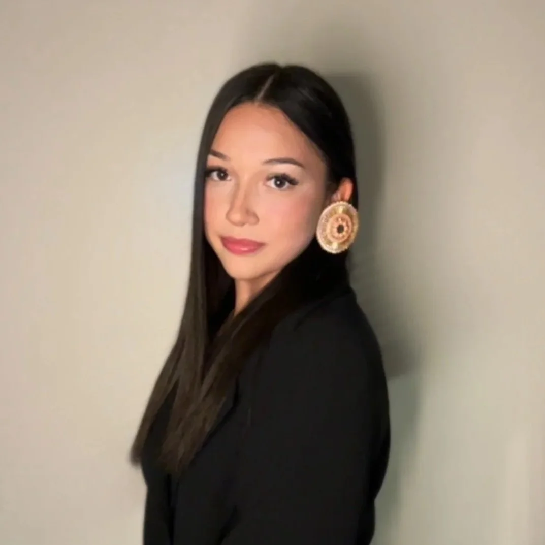 A young woman with long dark hair, wearing large circular earrings, a black top, and standing against a plain background.