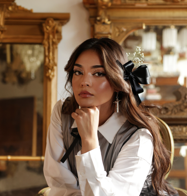 A young woman with long brown hair, wearing a white blouse and a black ribbon hair accessory, sitting in an ornate room with gilded mirrors and antique furniture.