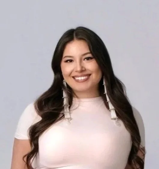 A young woman with long dark hair, smiling, wearing a white top and long earrings, against a plain gray background.