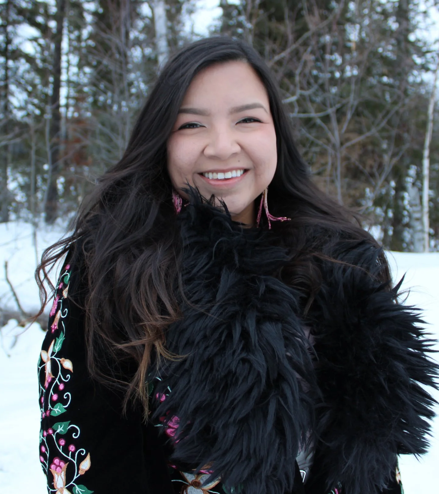 A woman smiling outdoors in winter, wearing a black coat with colorful embroidery and a black feathered scarf, with snow and trees in the background.