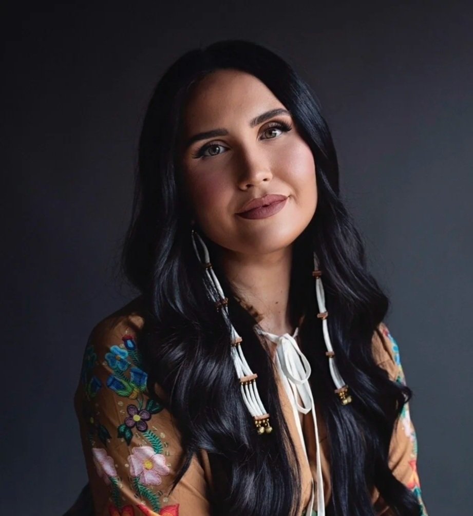 A woman with long, wavy black hair and light skin, wearing a colorful embroidered top and beaded earrings, looking at the camera with a slight smile against a dark background.