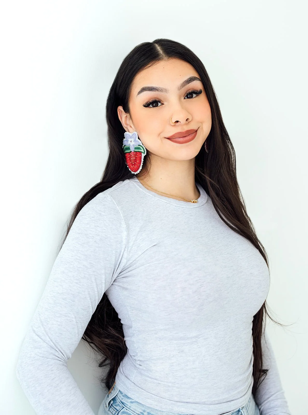 A young woman with long dark hair wearing a gray long-sleeve top, large strawberry-shaped earrings with floral accents, and a small necklace, standing against a white background, smiling at the camera.