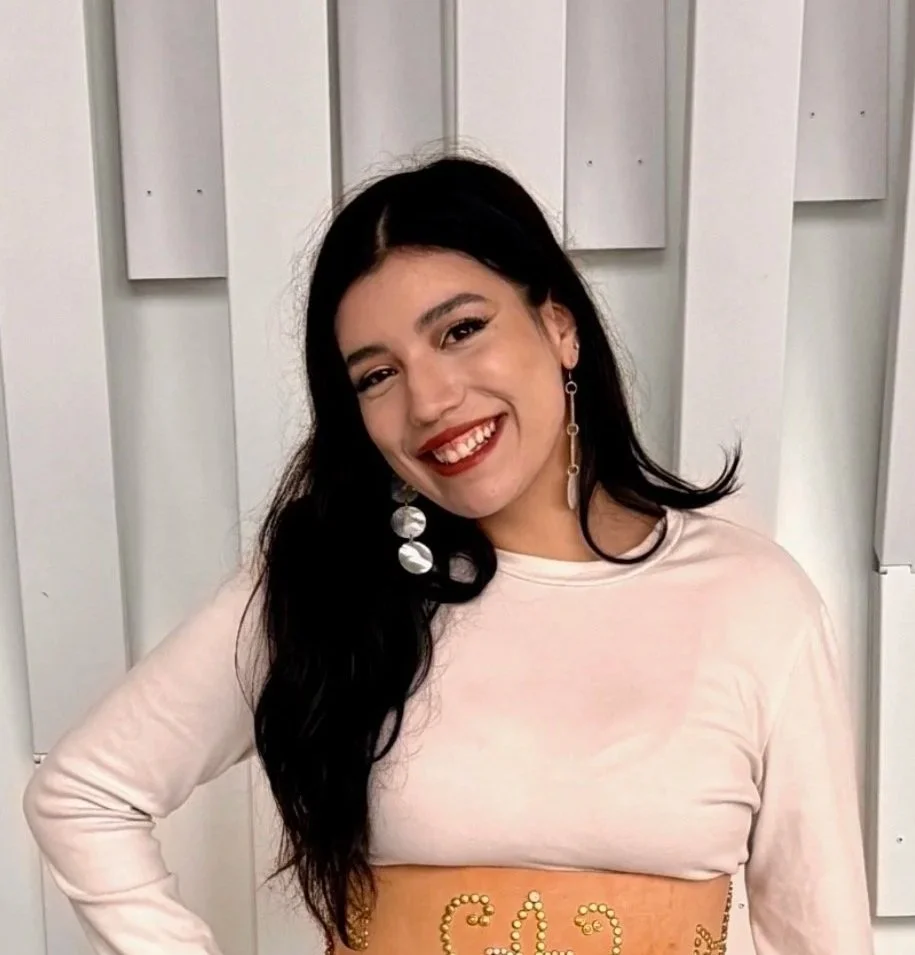 Young woman with long black hair and earrings, smiling, wearing a light pink top and a beige skirt with gold decorative patterns, standing against a white wall with vertical panels.