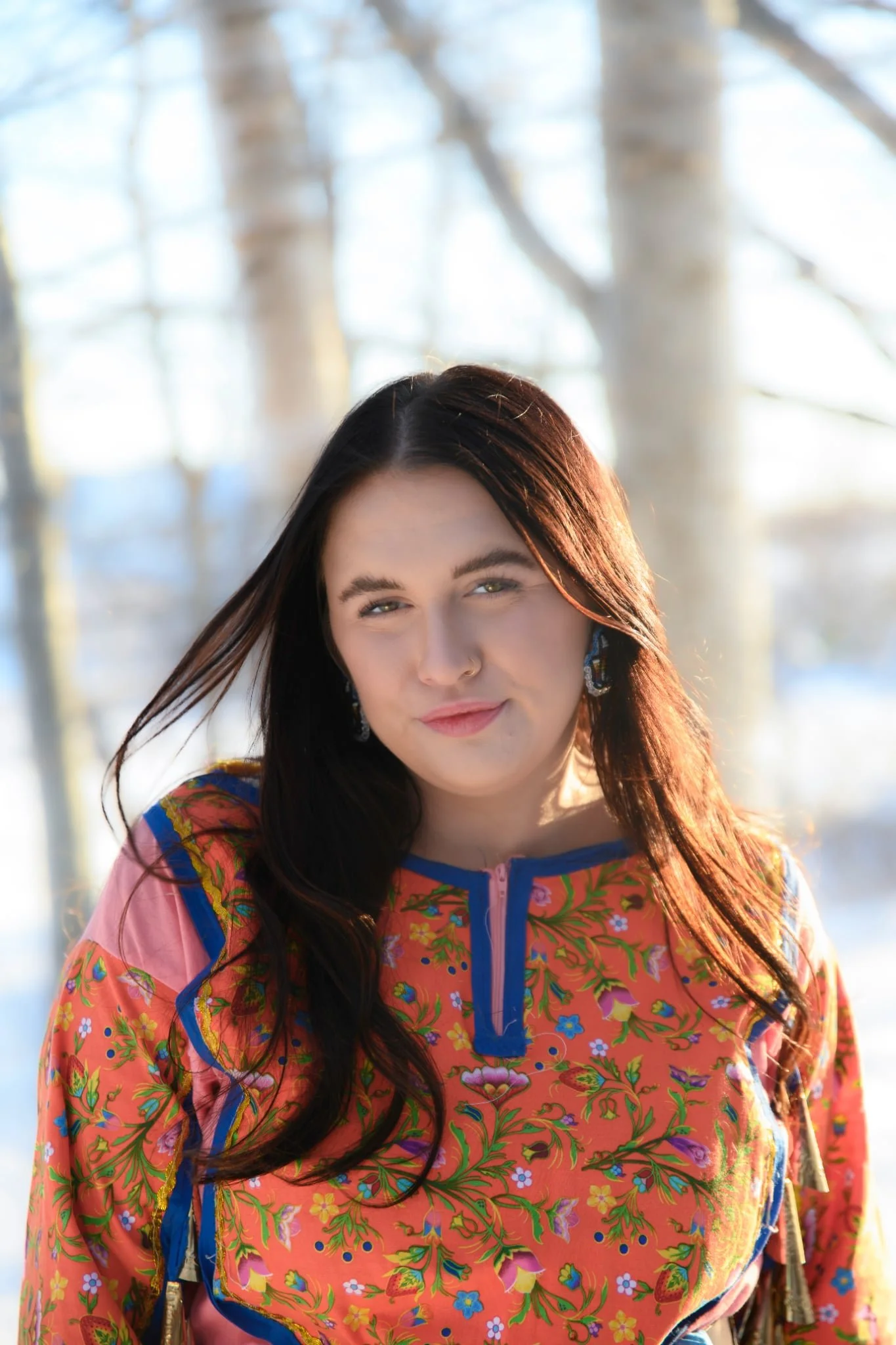 A woman with long dark hair and light skin, wearing a colorful floral jacket, standing outdoors with trees in the background during daylight.