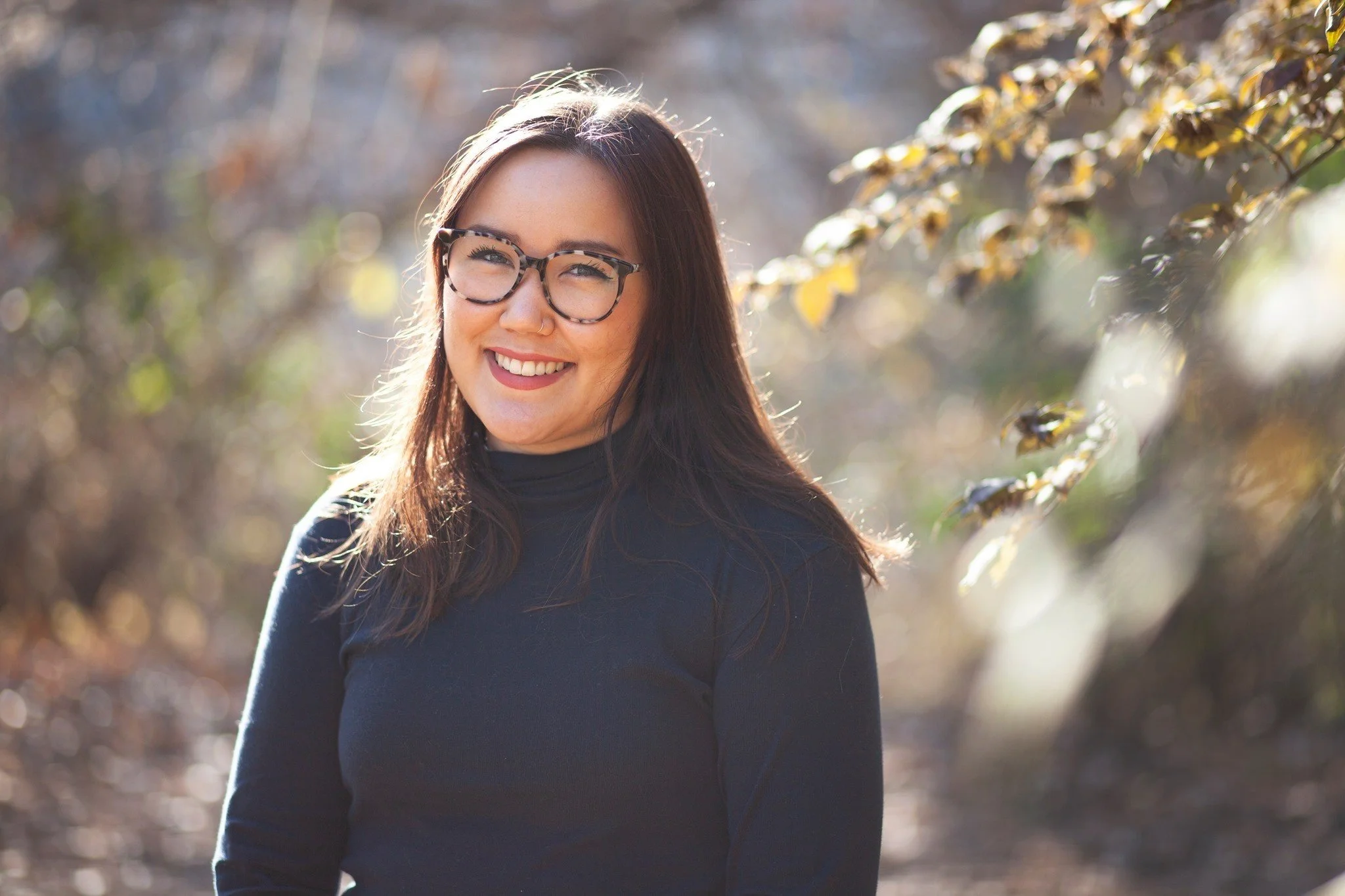 A young woman with long dark hair, wearing glasses and a black turtleneck, smiling outdoors with blurred autumn trees in the background.
