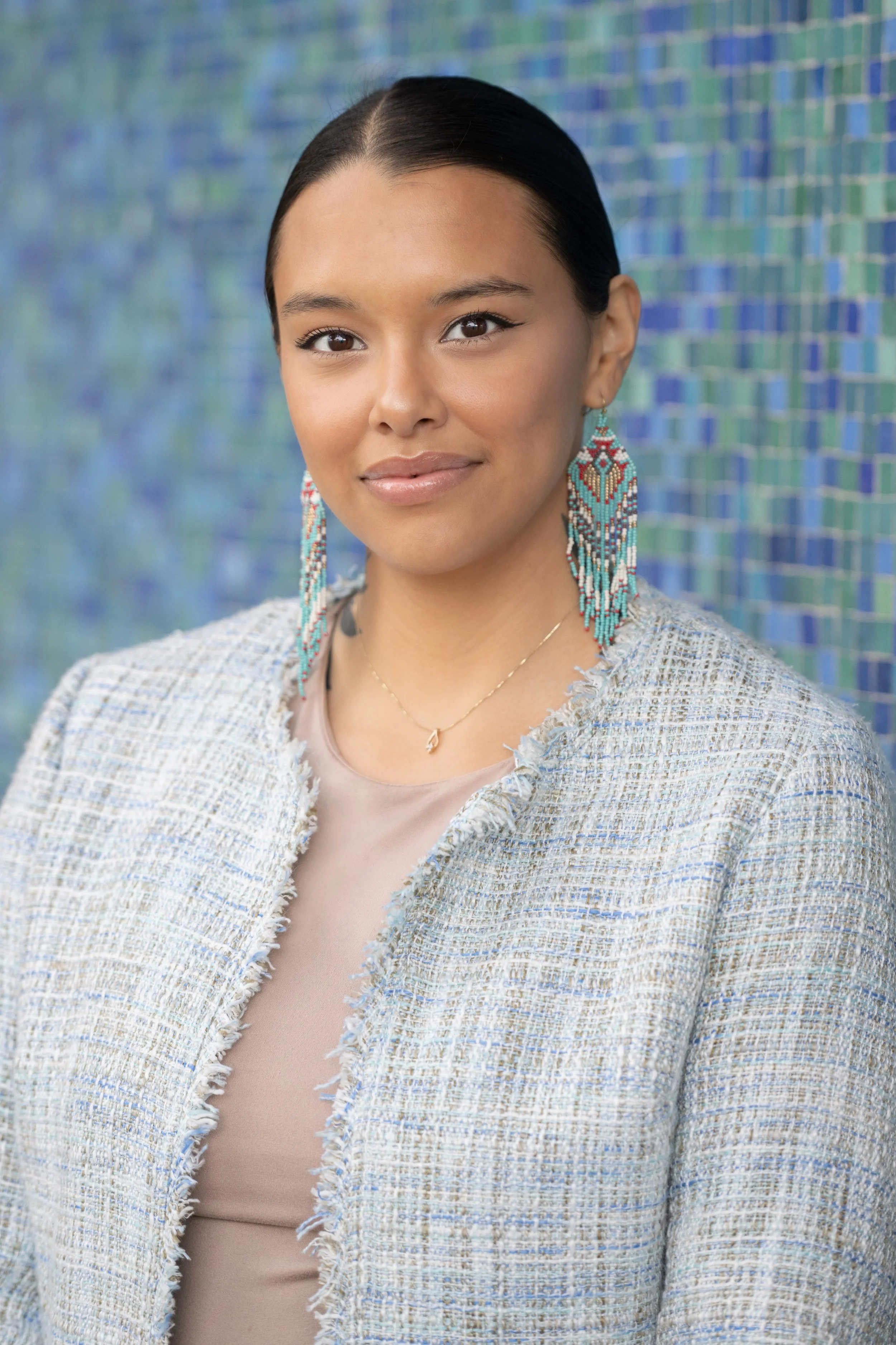 Woman with dark hair styled straight, wearing beaded earrings, a beige top, and a textured light-colored blazer, standing in front of a blue mosaic tile background.