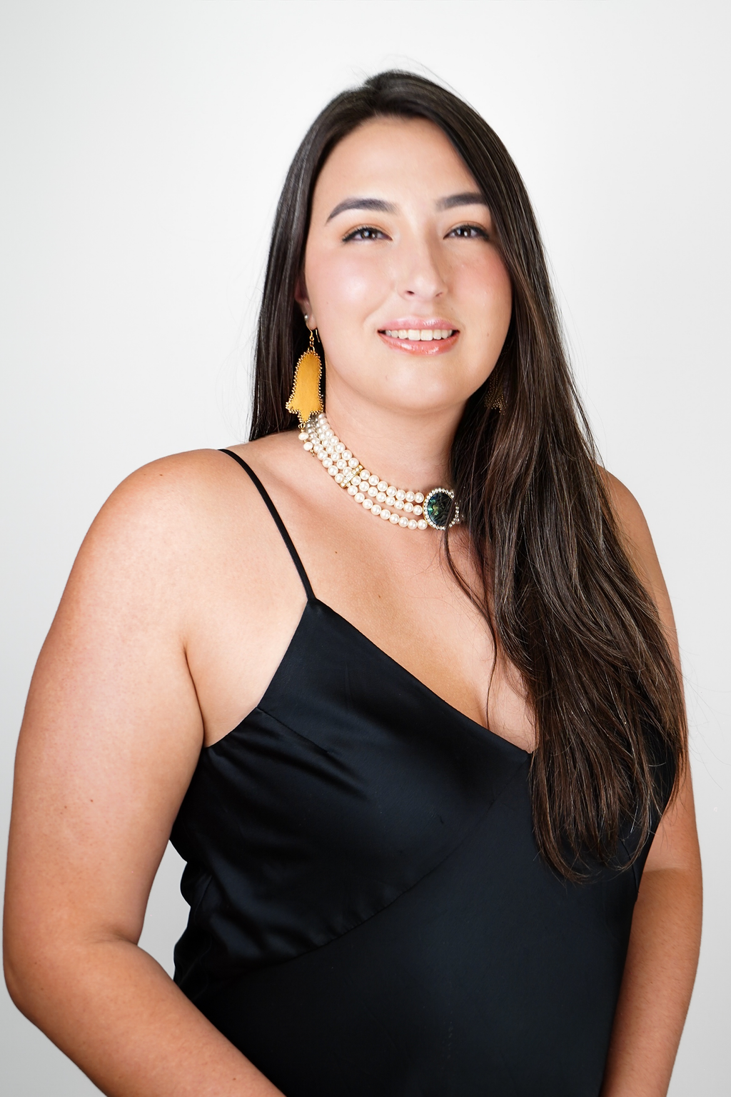 A woman with long dark hair wearing a sleeveless black top and purple flower earrings, standing in front of lush green ferns.