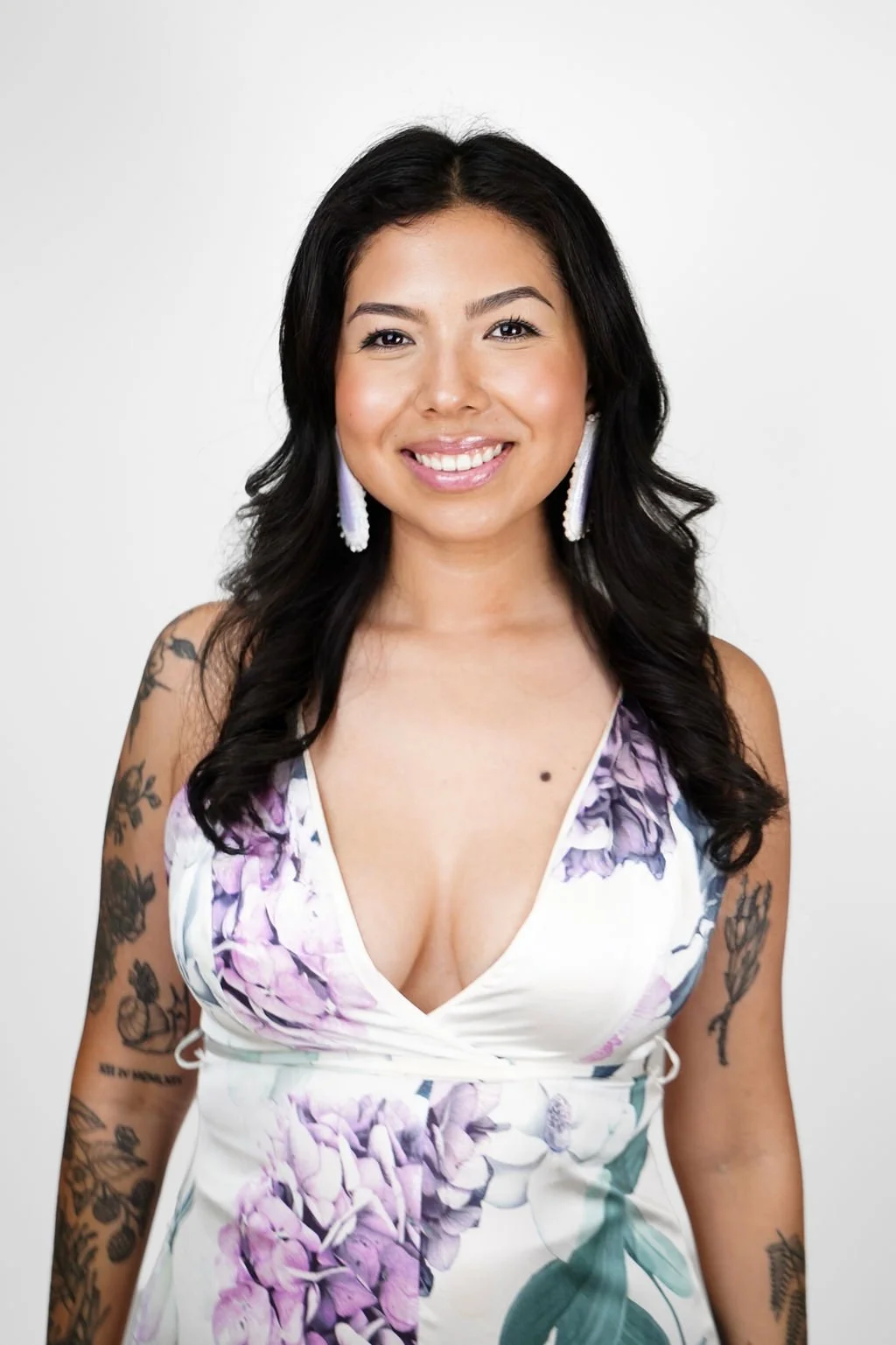A young woman with black wavy hair, adorned with red butterflies, smiling and wearing a white floral dress.