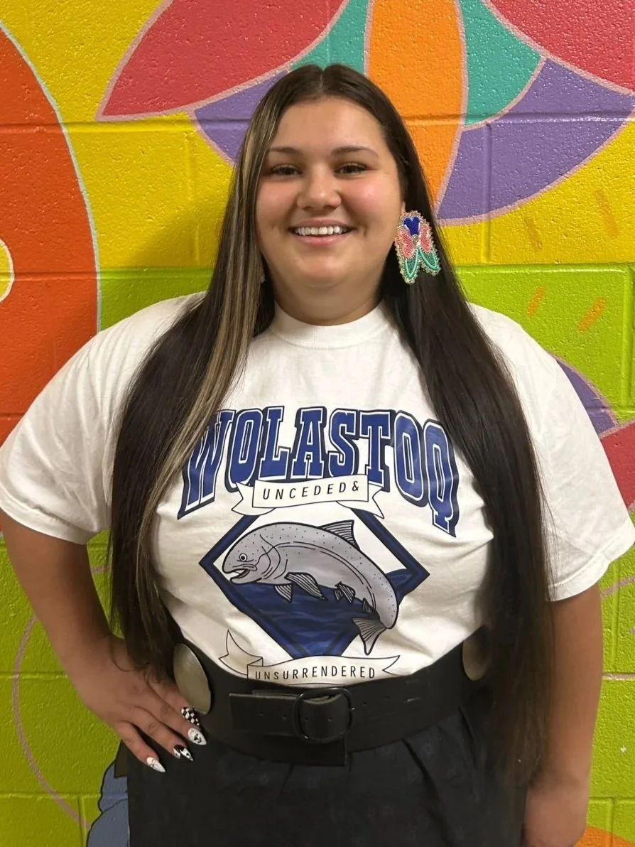 A smiling young woman with long dark hair, wearing a white T-shirt with a fish graphic and the words "WOLASTQ UNCEDDED & UNSURRENDERED," standing in front of a colorful mural with bright geometric shapes.