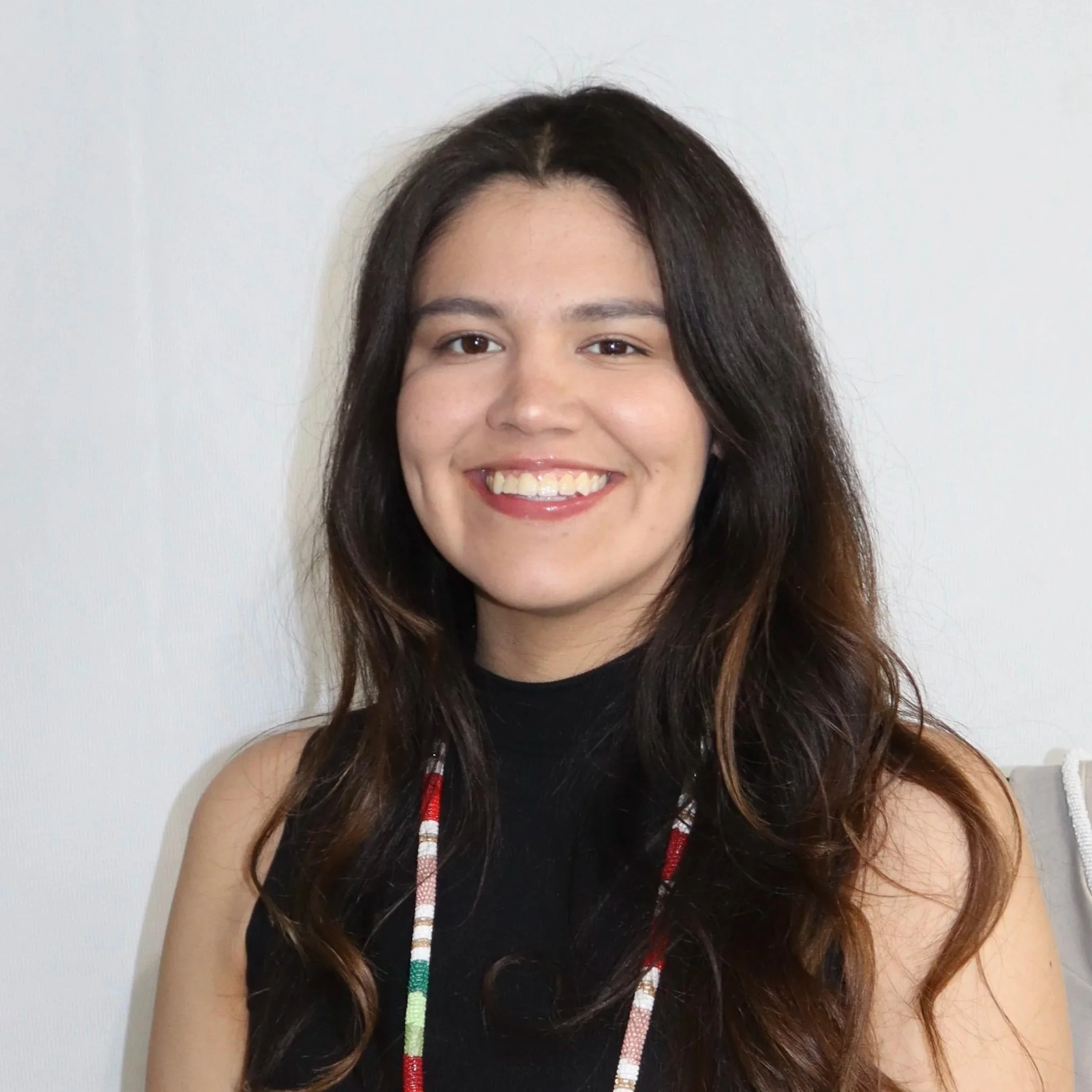 A young woman with long dark wavy hair, smiling, wearing a black sleeveless top and a colorful beaded necklace, sitting against a plain white wall.