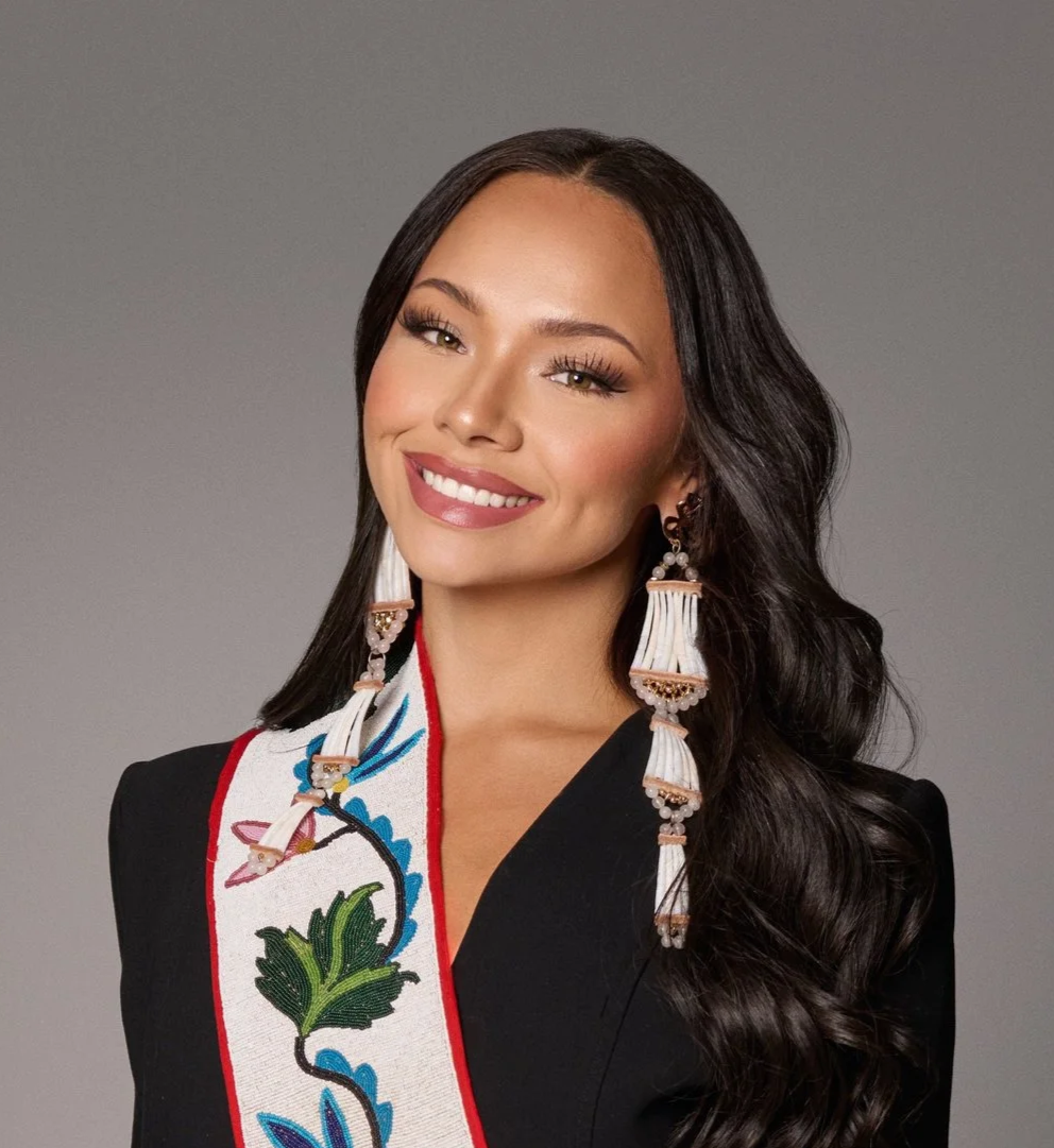 A woman with long, dark, wavy hair, smiling, wearing a black blazer, large white beaded earrings with pink accents, and a sash with colorful floral embroidery.