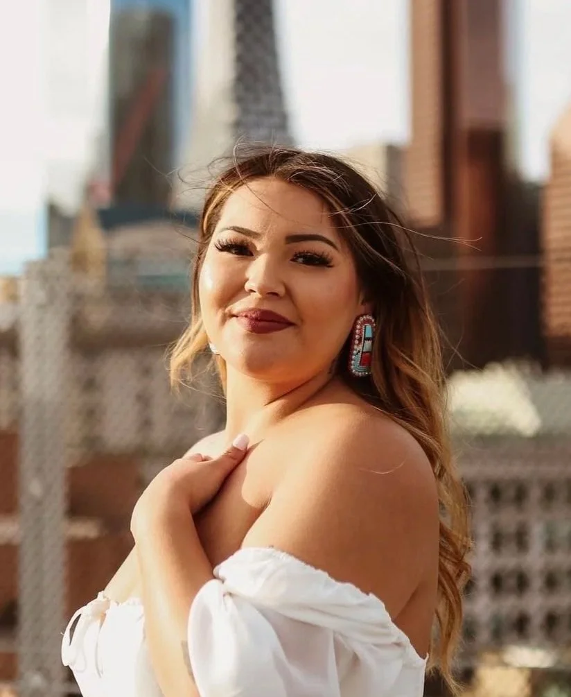 A woman with long, wavy hair and makeup, wearing a white off-the-shoulder top and colorful earrings, standing outdoors with a blurred cityscape in the background, smiling at the camera.