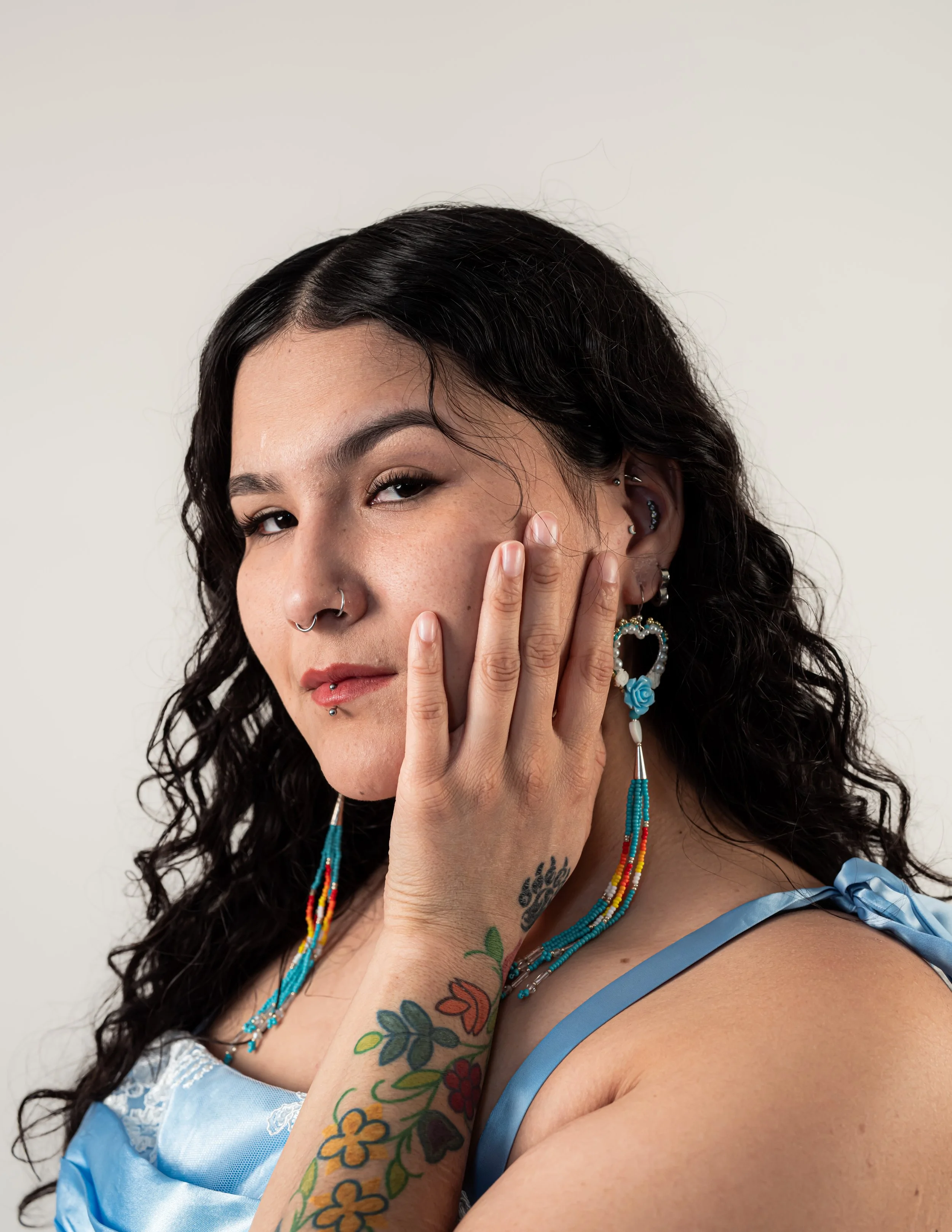 Close-up portrait of a woman with dark curly hair, multiple facial piercings, colorful tattoos on her arm, and wearing blue jewelry and a blue satin dress.