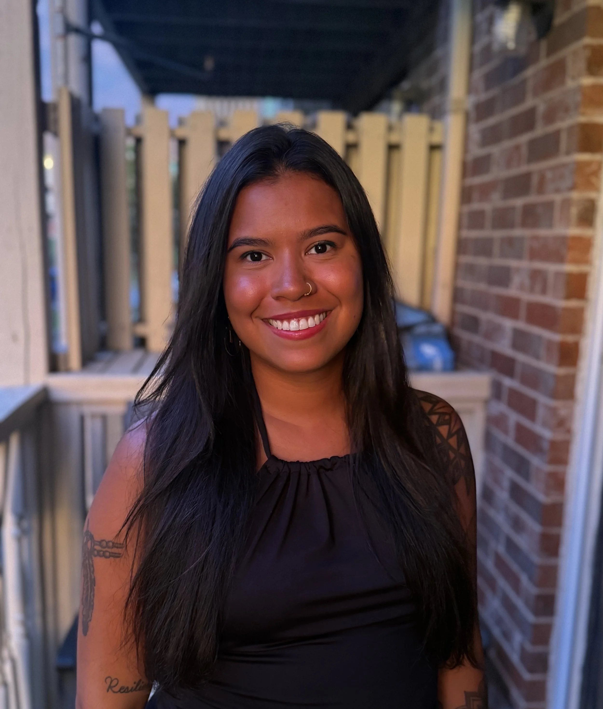 A young woman with long black hair, wearing a black sleeveless top, standing outdoors on a porch, smiling at the camera.