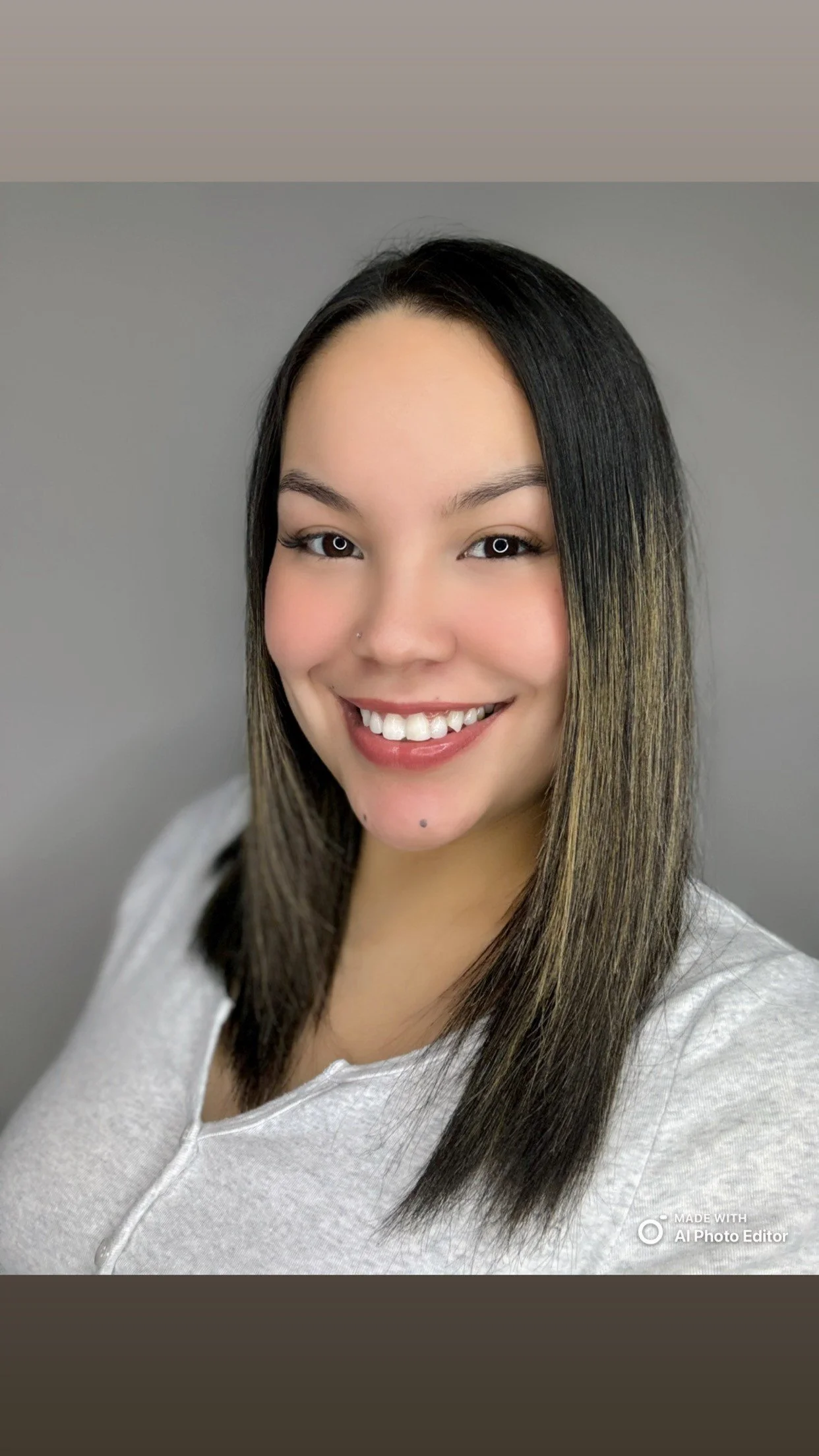 Portrait of a young woman with straight dark hair and a nose piercing, smiling and wearing a light-colored top, against a plain gray background.