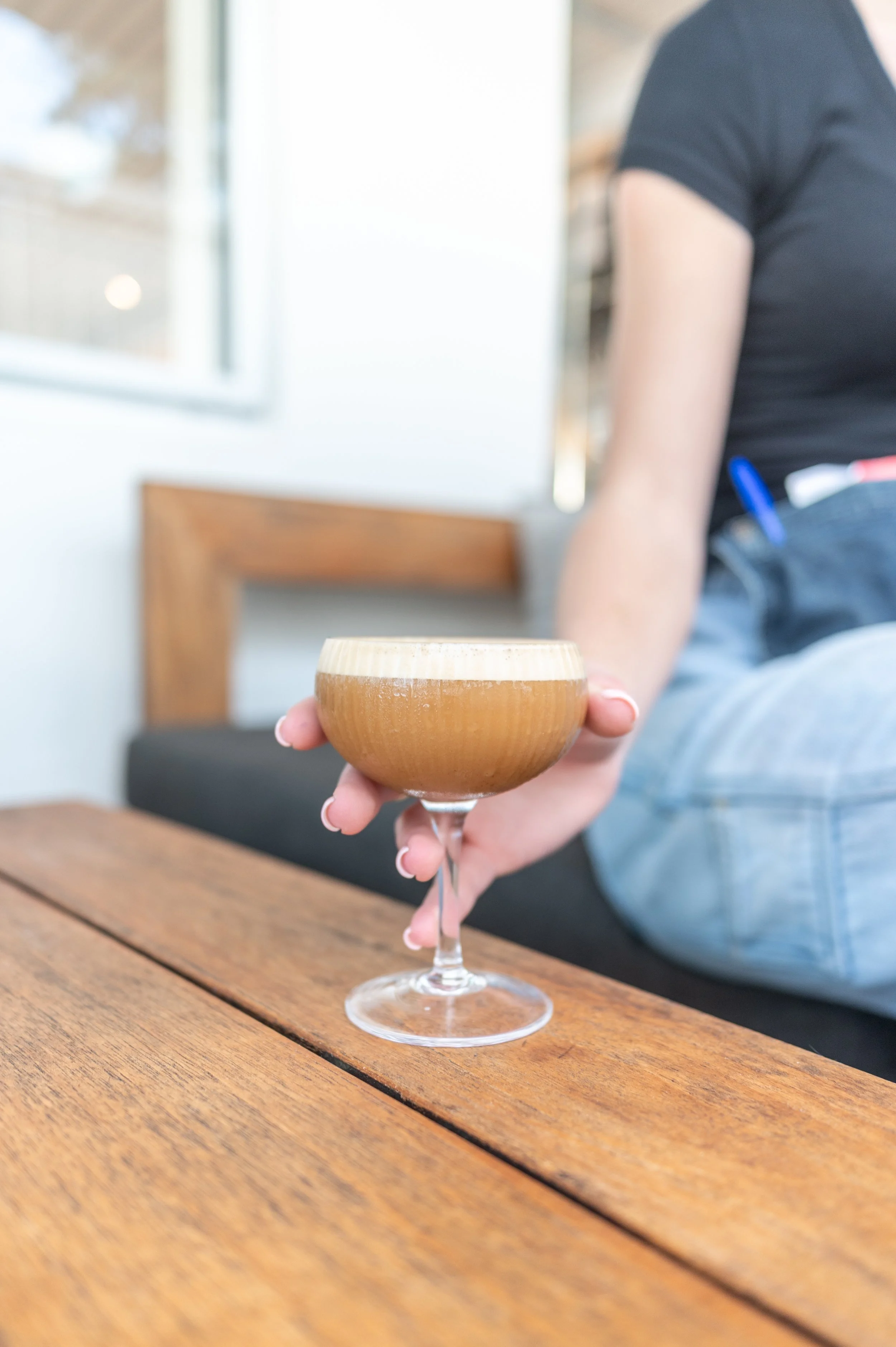 Person sitting on a bench offering a cocktail in a coupe glass on a wooden table, wearing a black shirt and jeans.
