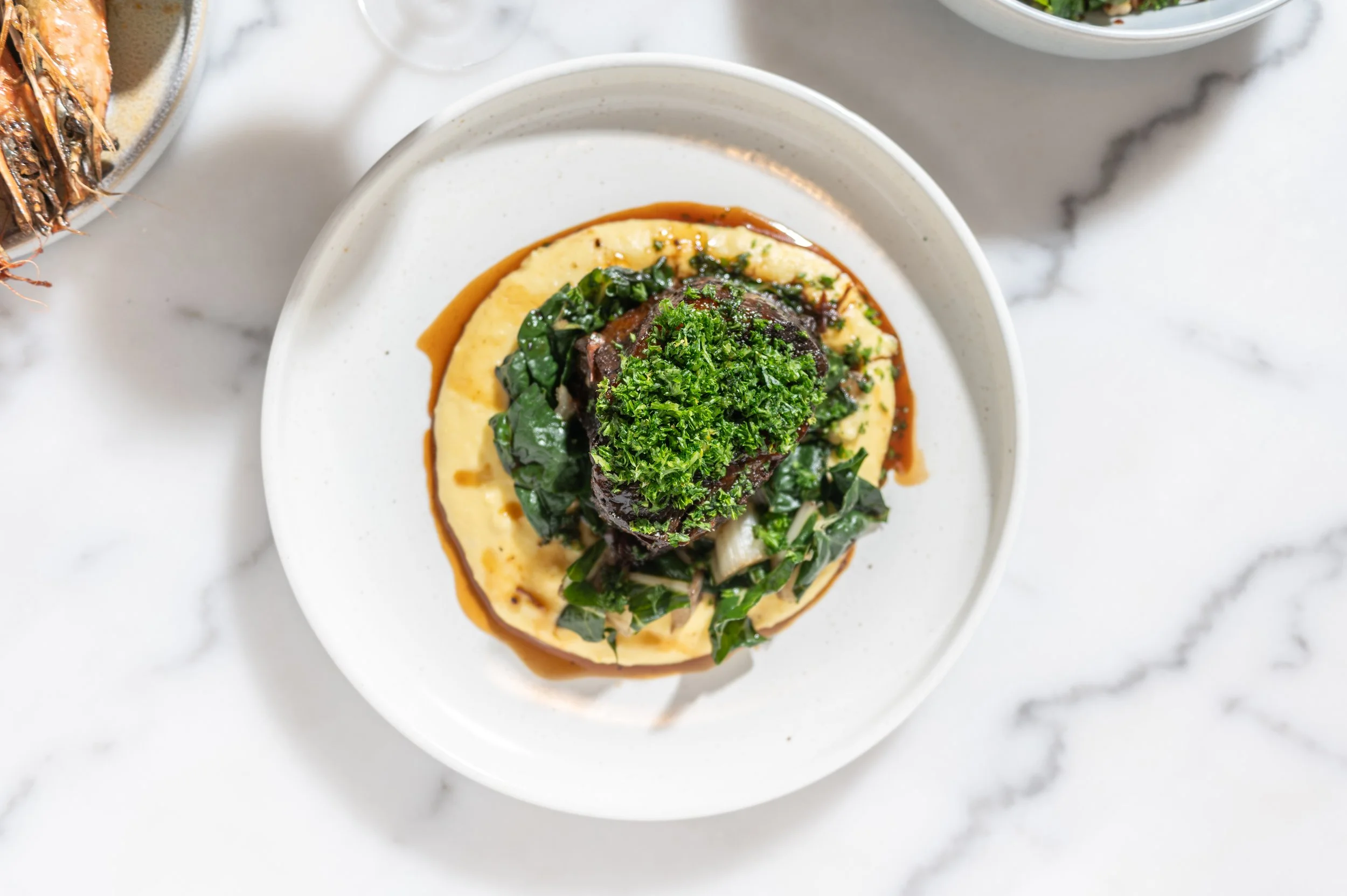 A white bowl filled with mashed potatoes topped with a serving of braised beef, sautéed greens, and garnished with fresh parsley, with additional dishes partially visible to the sides on a white marble surface.