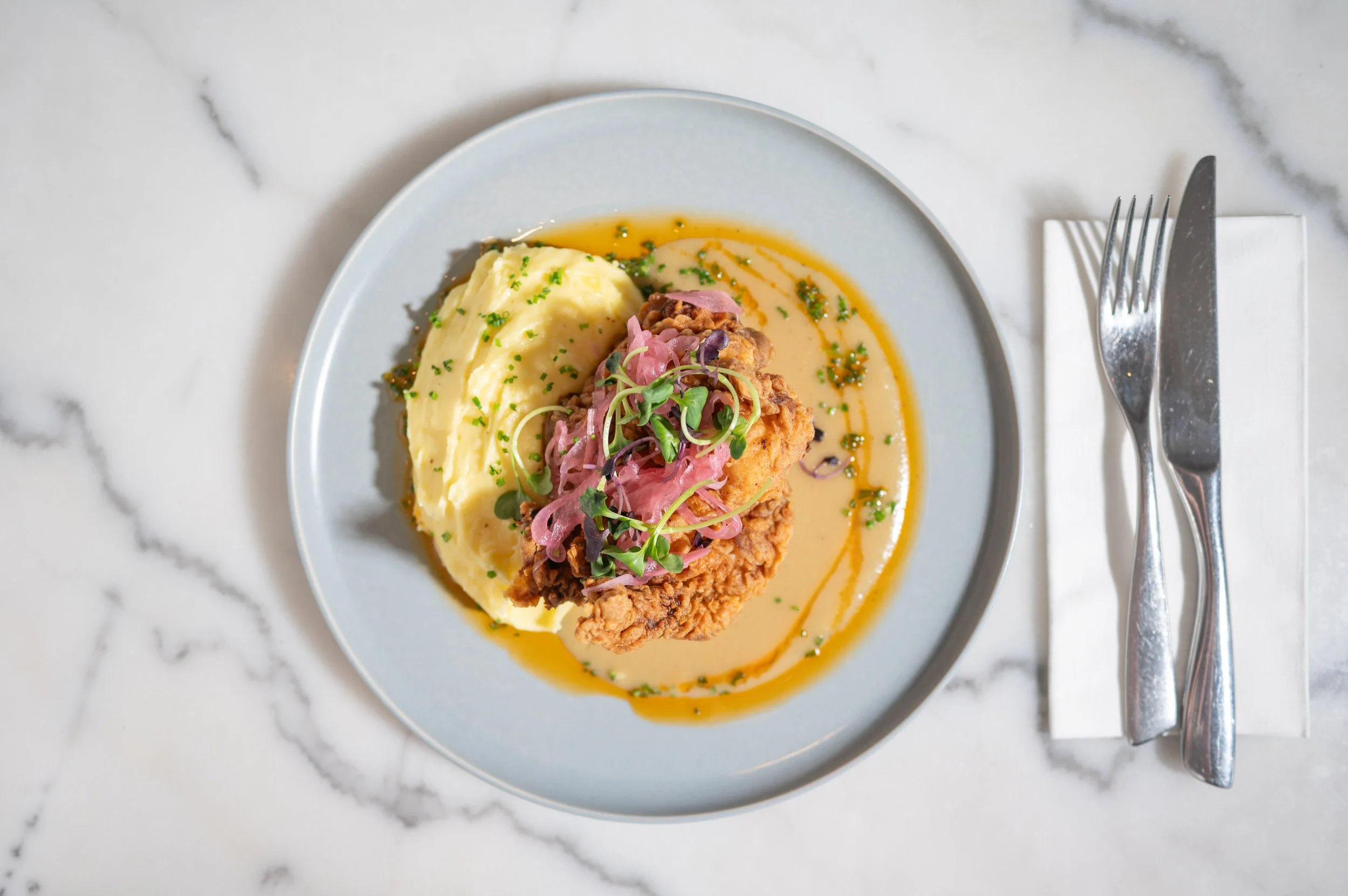 Plate of mashed potatoes with herbs, a fried chicken cutlet topped with pickled onions and microgreens, garnished with sauce and herbs, served on a light blue plate on a marble table.