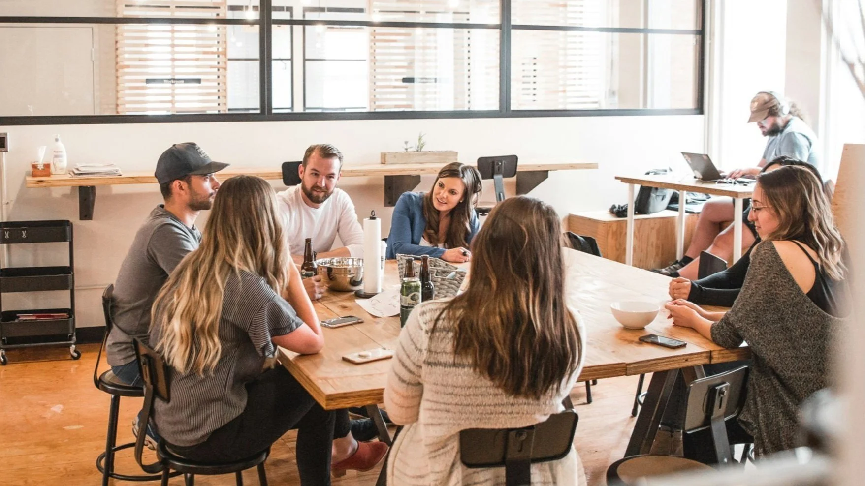 Group of people having a meeting around a wooden table in a modern office, some with drinks and notebooks, with one person working on a laptop in the background.