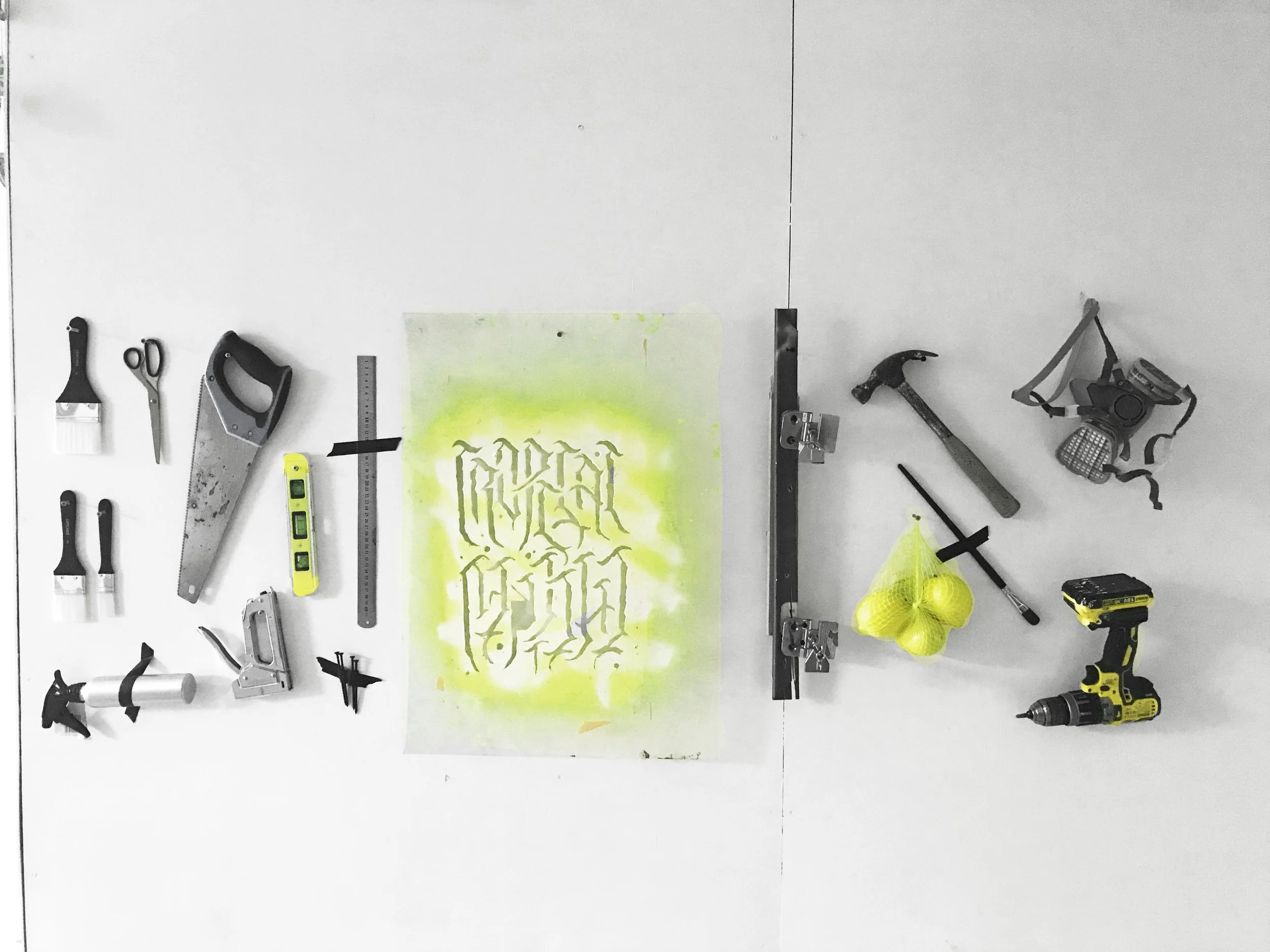 Tools, spray paint, a level, a saw, a hammer, a flashlight, a drill, a mask, and yellow onions on a white surface with a green spray-painted paper with illegible black graffiti-style writing.