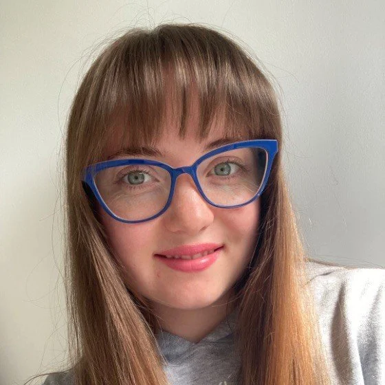 Woman with curly hair wearing a teal dress and a leaf-shaped necklace, smiling against a dark background.