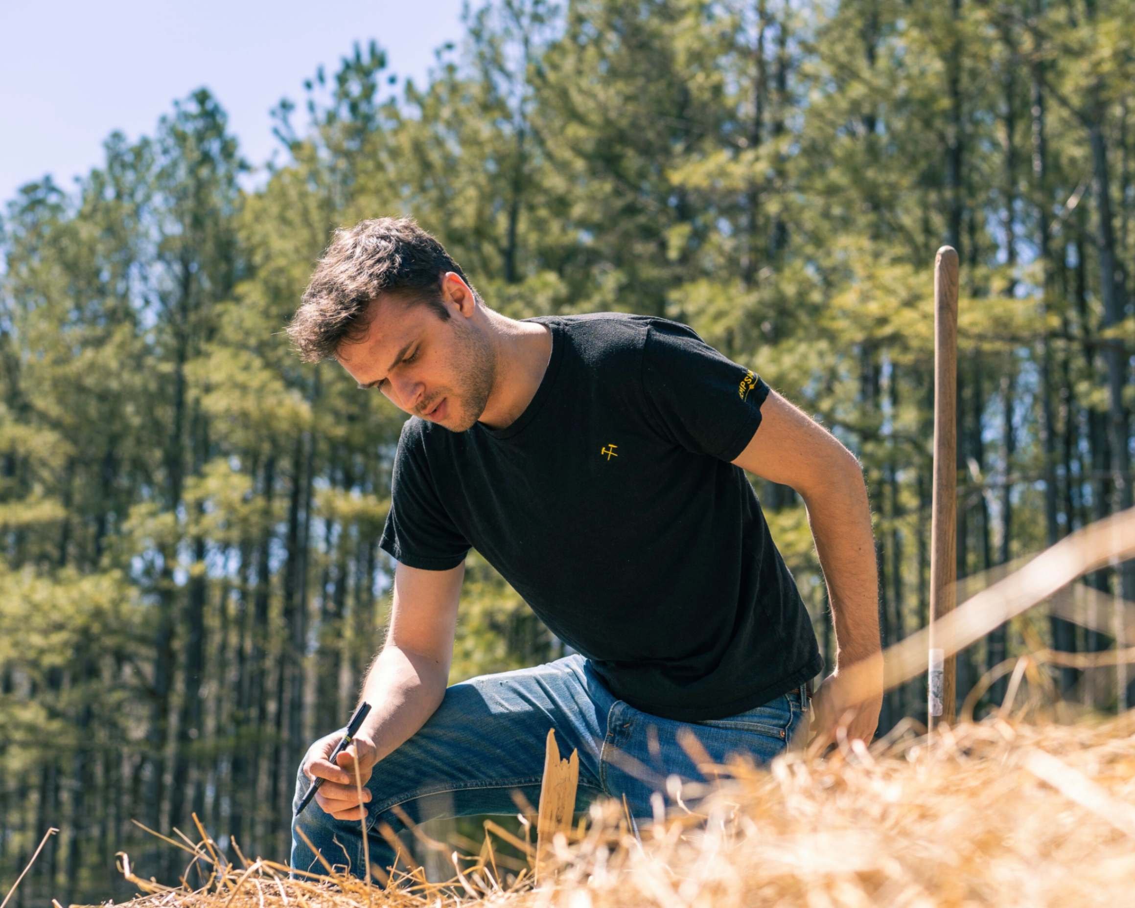 Man in a black hemp shirt with gold embroidery working in a field with trees behind him.