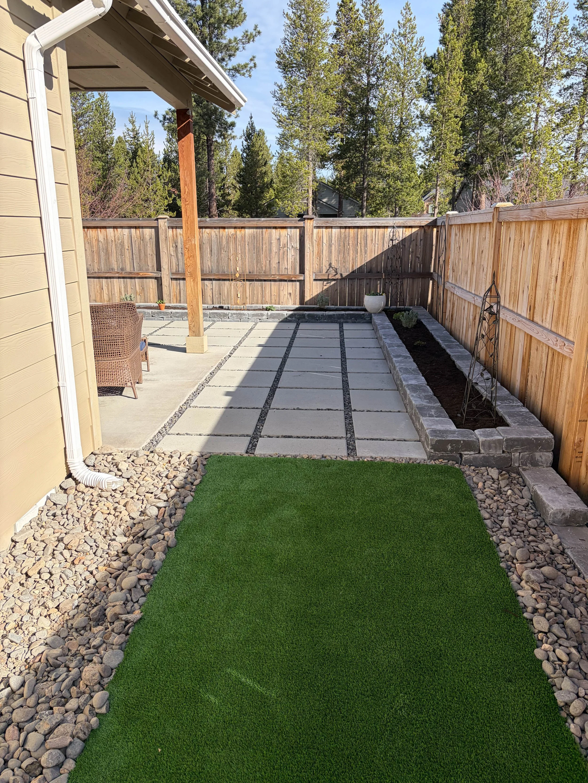 Backyard garden with a small patch of artificial grass, a gravel border, a patio with concrete pavers, and a raised brick garden bed with plants, enclosed by a wooden fence.
