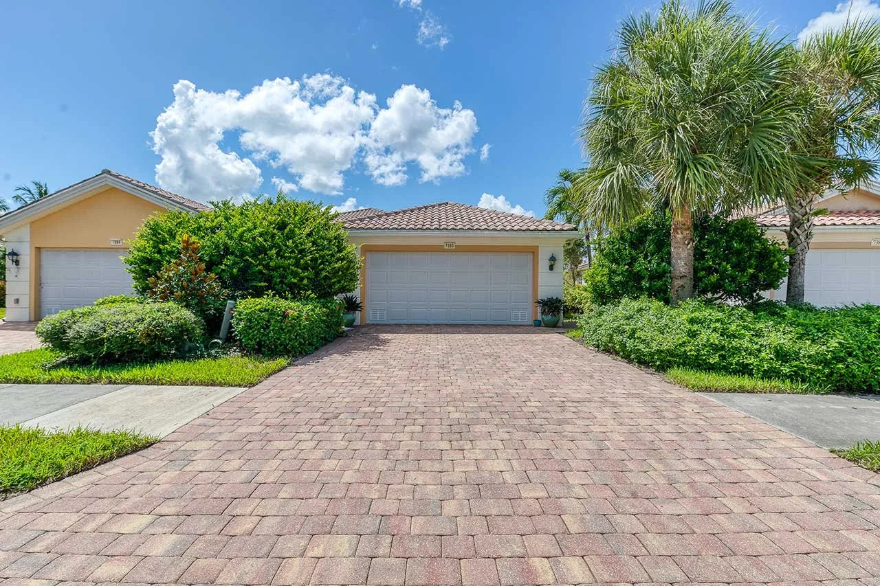A residential neighborhood with three single-family homes, each with a tile roof and white garage doors, surrounded by lush greenery, palm trees, and a brick driveway under a partly cloudy blue sky.