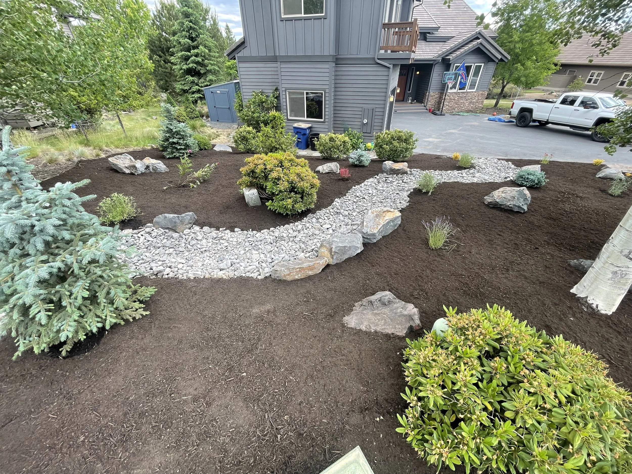 A landscaped front yard with freshly planted shrubs, a stone pathway, and dark mulch around the plants, in a suburban neighborhood.