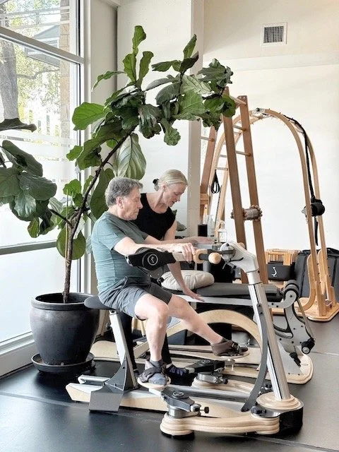 Older man and woman exercising on a rowing machine indoors near a large window and a tall potted plant.