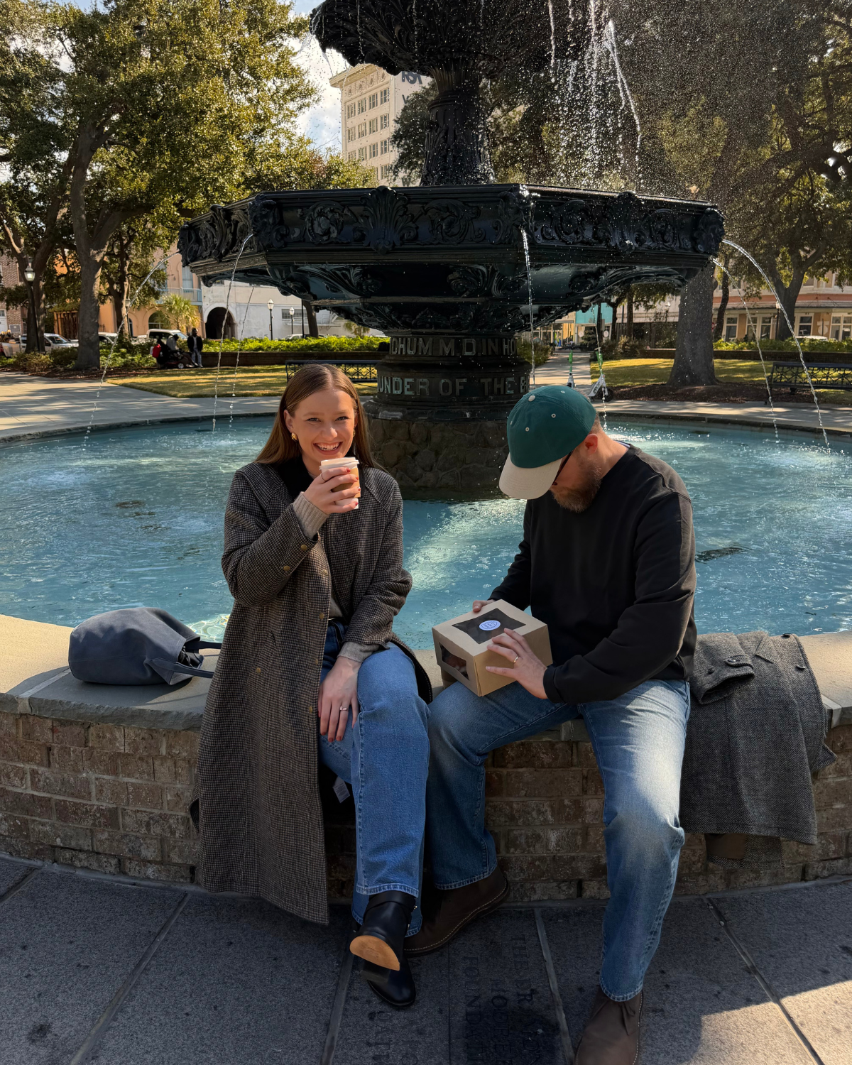 A woman and a man sitting by a fountain in a park, the woman is holding a drink and smiling while the man is looking into a box, with trees and buildings in the background.