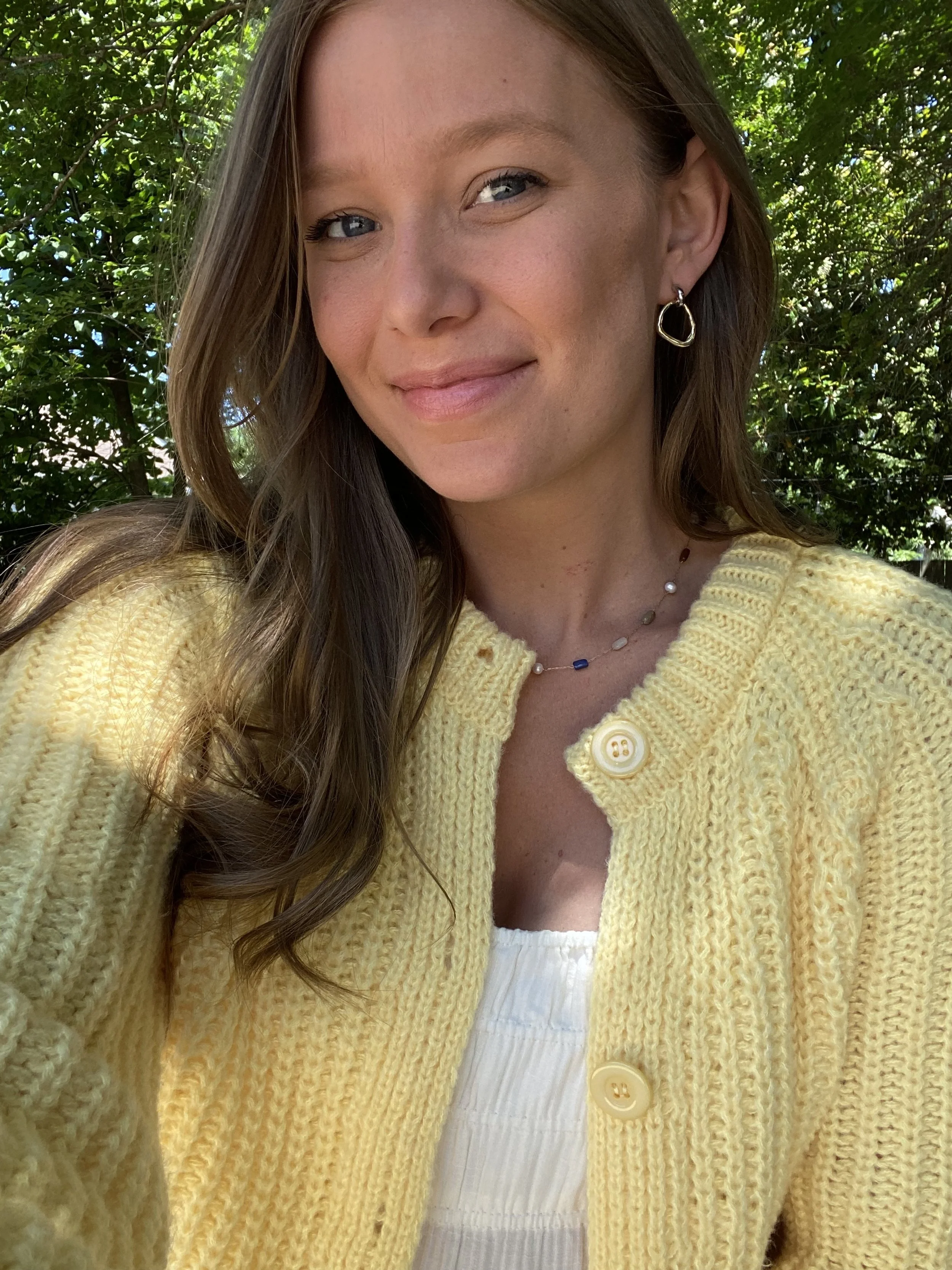 A woman smiling outdoors, wearing a yellow knit cardigan, white top, earrings, and a necklace, with trees in the background.