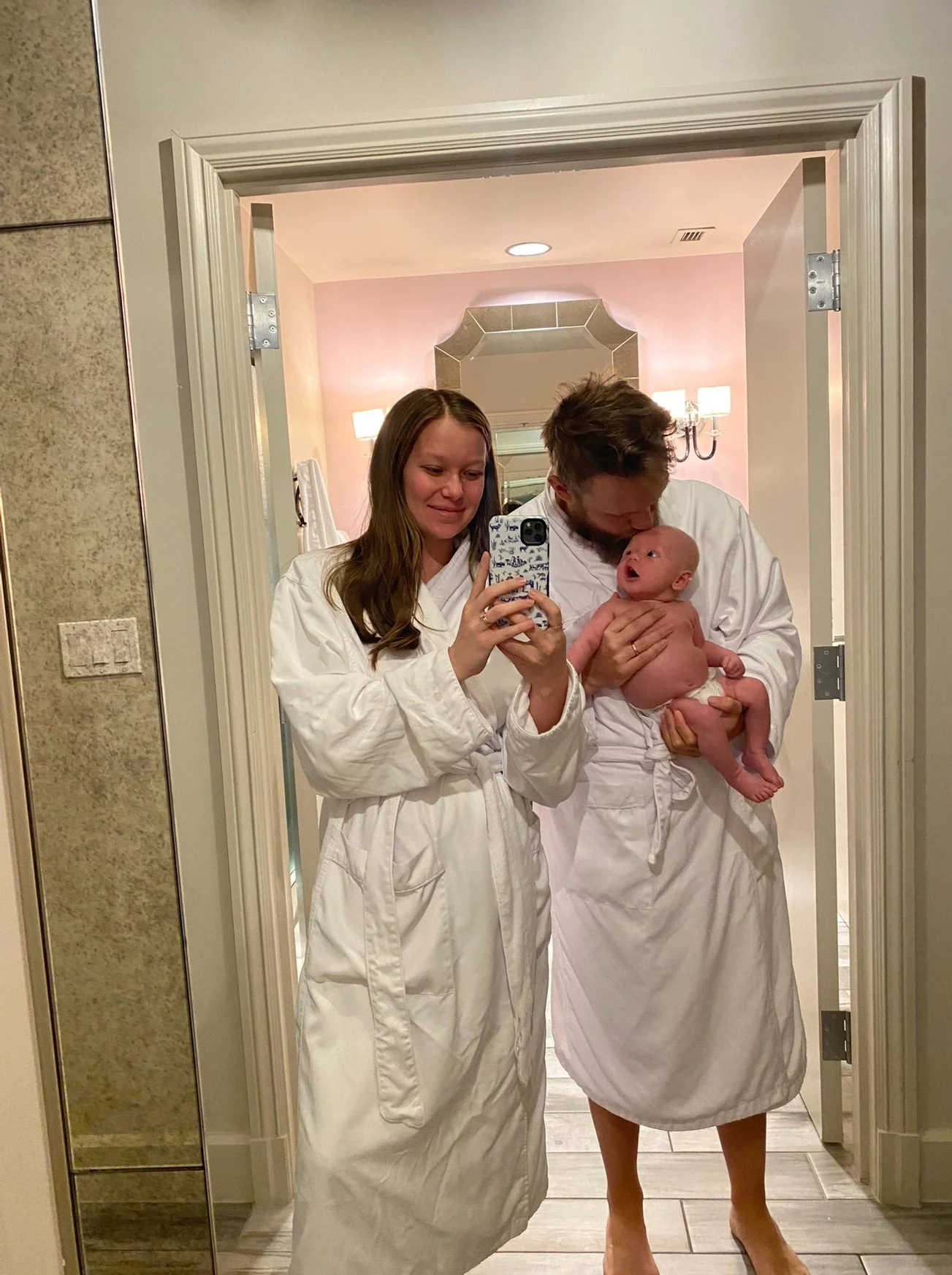 A woman and a man in white robes taking a mirror selfie in a bathroom. The woman is smiling while the man holds a newborn baby close, kissing the baby's head.