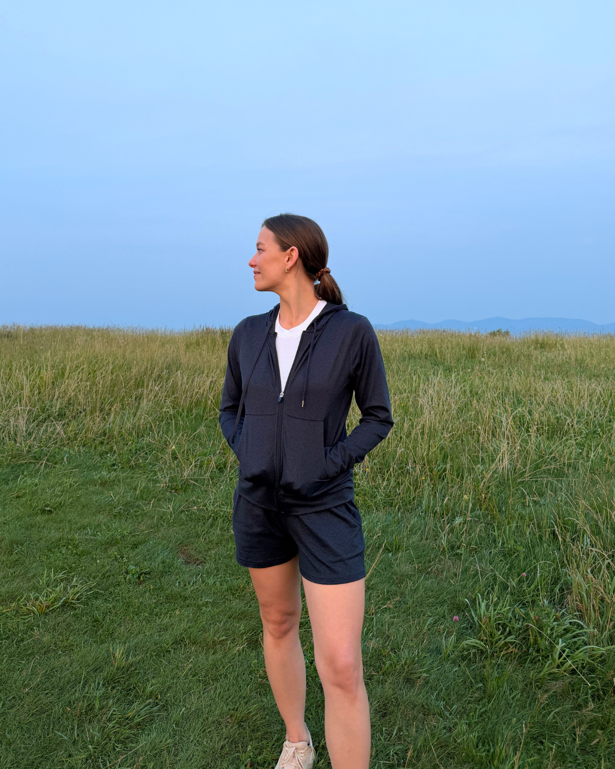 Woman in black athletic jacket and shorts standing in a grassy field with hills in the background during daytime.