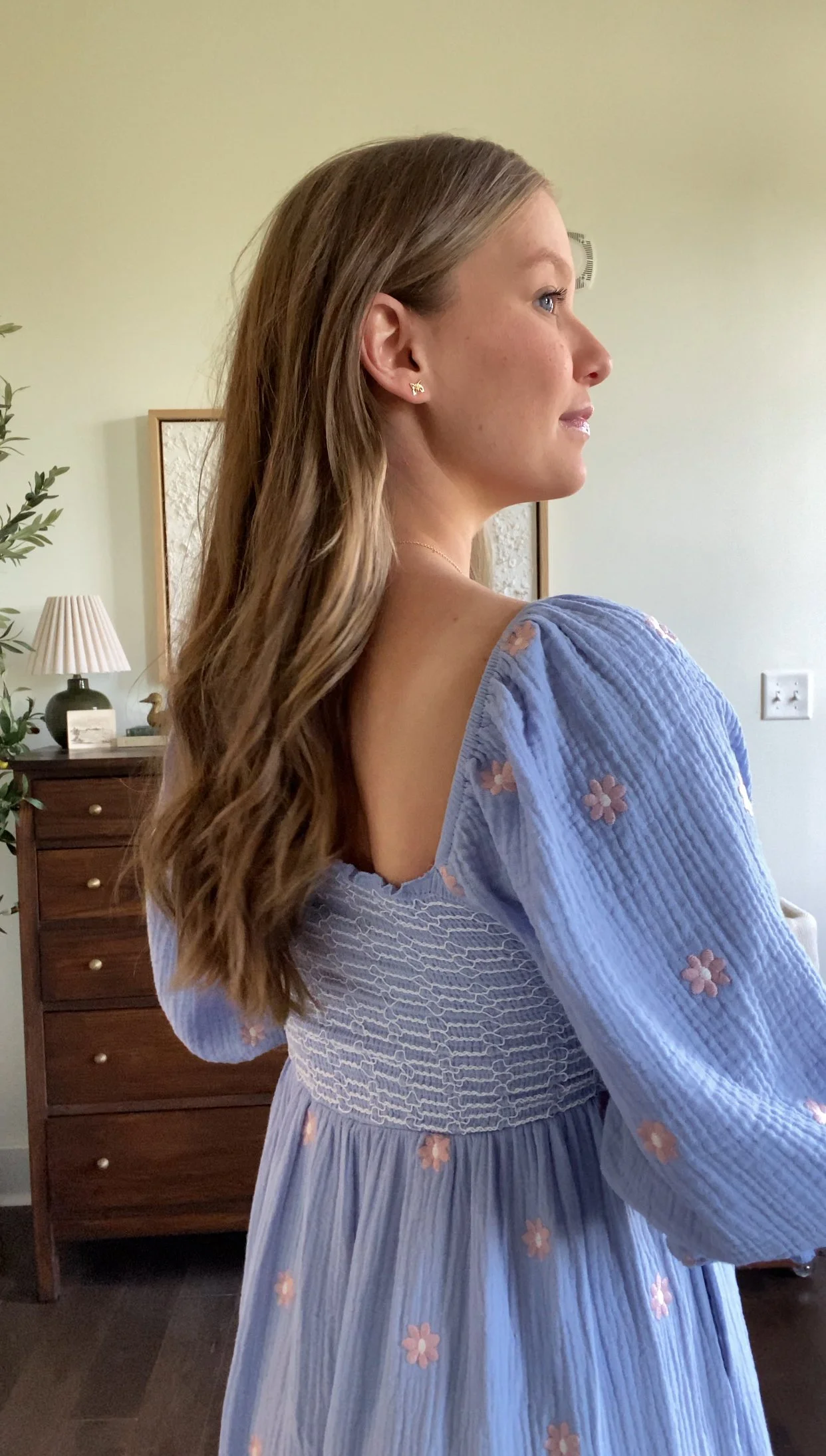 Profile view of a woman with long wavy hair wearing a light purple dress with floral embroidery in a room with light green walls, a wooden dresser, a table lamp, a small decorative figure, and greenery.