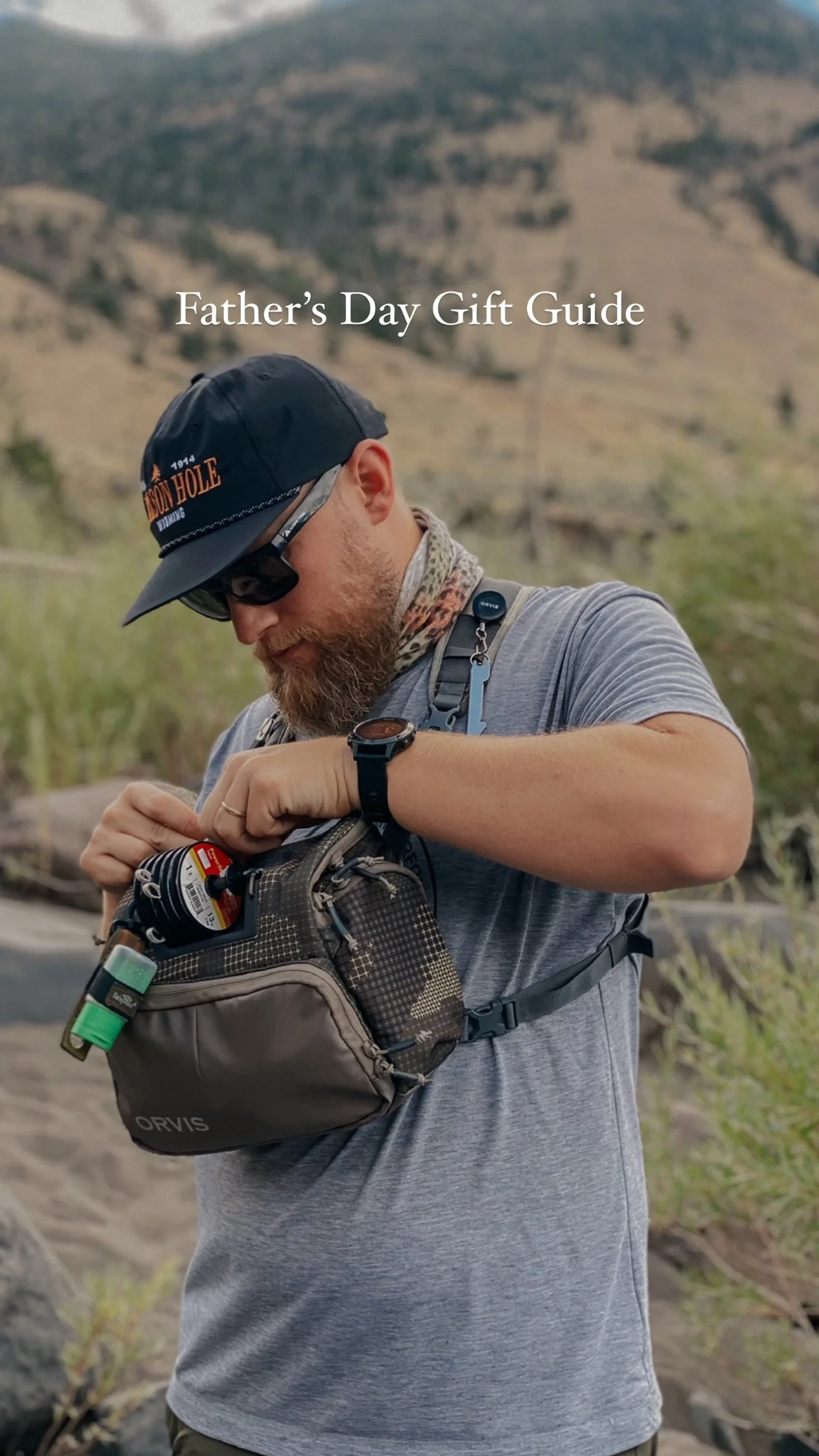 A man with a beard, wearing a baseball cap, sunglasses, and a gray t-shirt, is opening a small outdoor backpack, with a natural landscape of grass, rocks, and hills in the background, while preparing for an outdoor activity.
