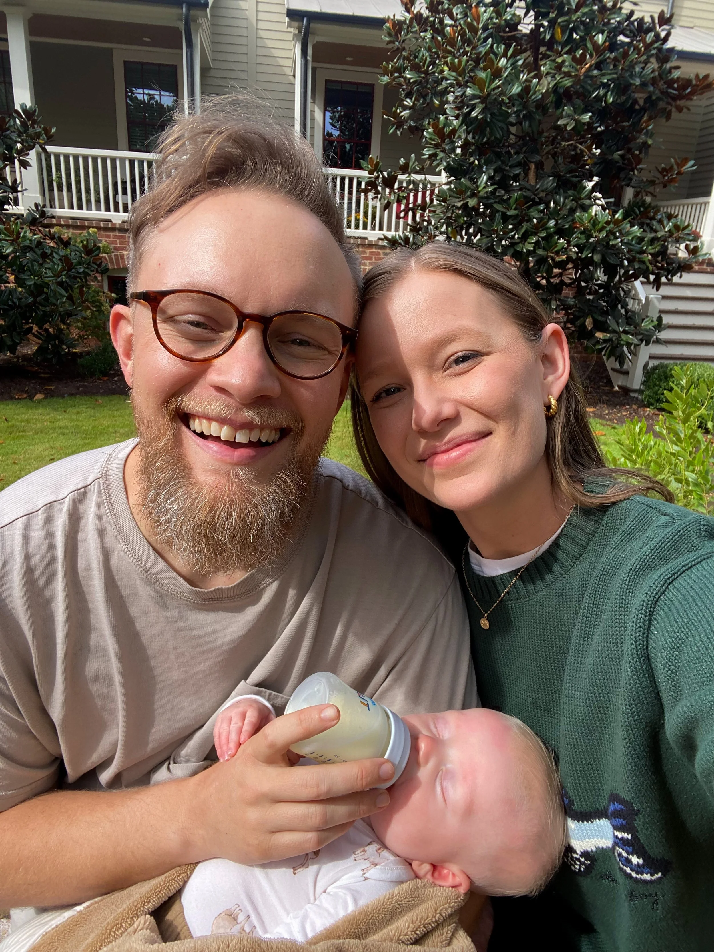 A smiling couple feeds a baby with a bottle in a backyard, with a house and trees in the background.