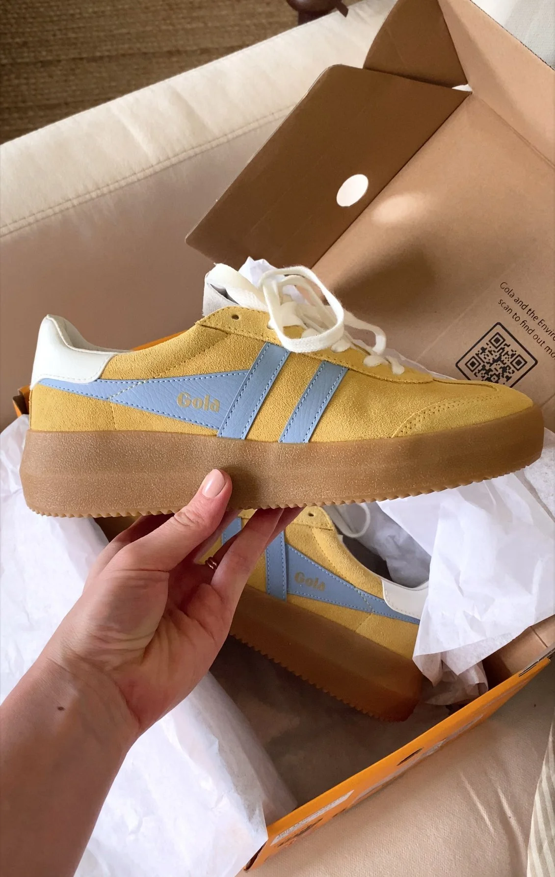 A yellow and light blue Gola sneaker with a brown sole, being held over its box with tissue paper inside.