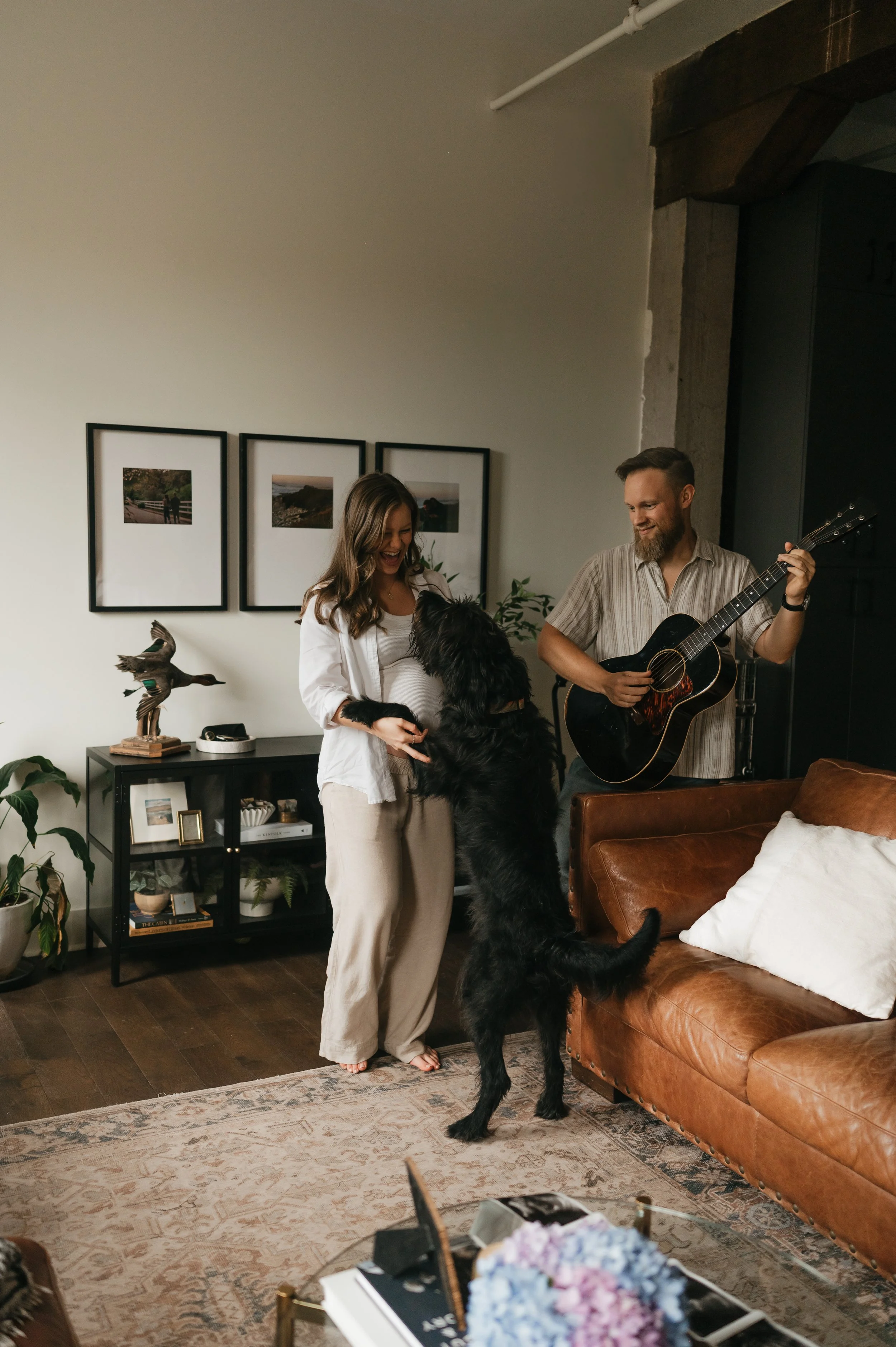 A woman and a man are inside a living room, with the woman smiling at a large black dog that is standing on its hind legs, touching her hands. The man is playing a guitar and smiling at the interaction. The room has framed photos on the wall, a dark-