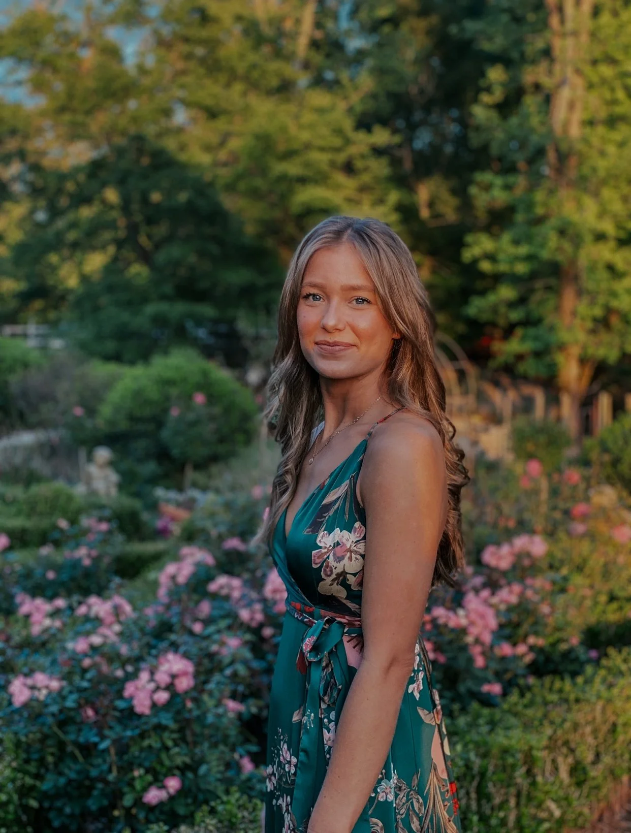 Woman in floral dress standing in garden with roses and trees in the background.