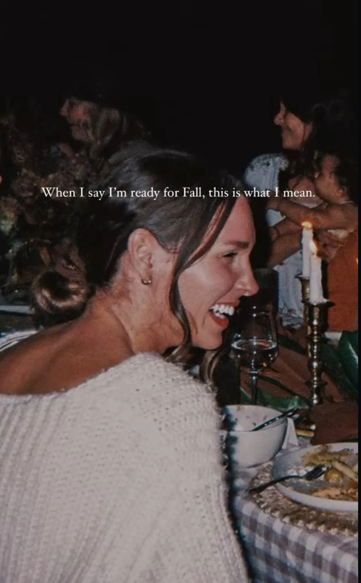 A woman with a joyful smile sitting at a dinner table with lit candles, a glass of white wine, and a bowl, with other people in the background at a nighttime gathering.