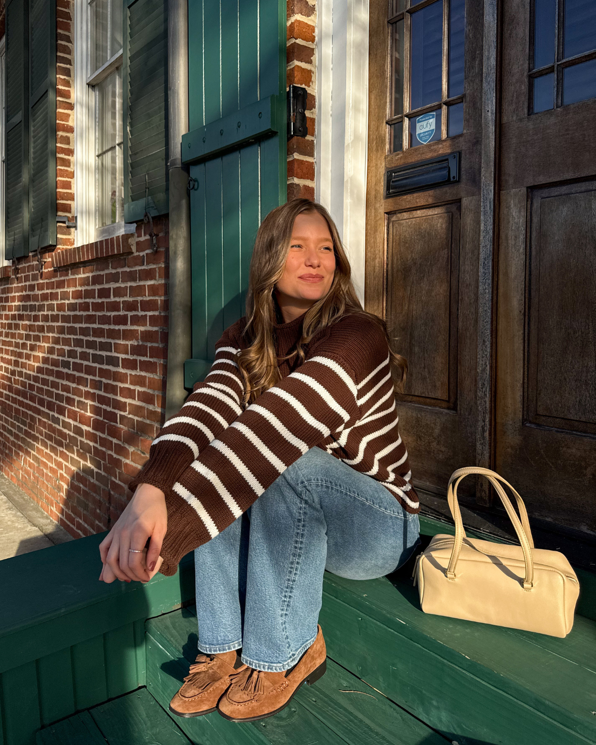 A woman sitting on a green wooden porch overlooking a brick house. She is wearing a brown and white striped sweater, blue jeans, and brown shoes. A yellow handbag is beside her.