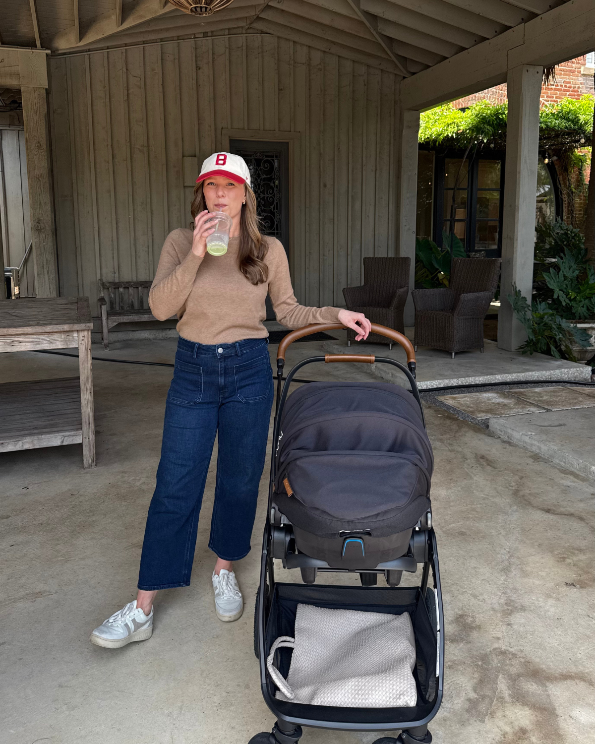 A woman with long brown hair wearing a white hat with a red B, a beige sweater, and blue jeans, drinking from a clear cup. She stands next to a black stroller with a white blanket inside, on an outdoor patio with wooden furniture and greenery in the 