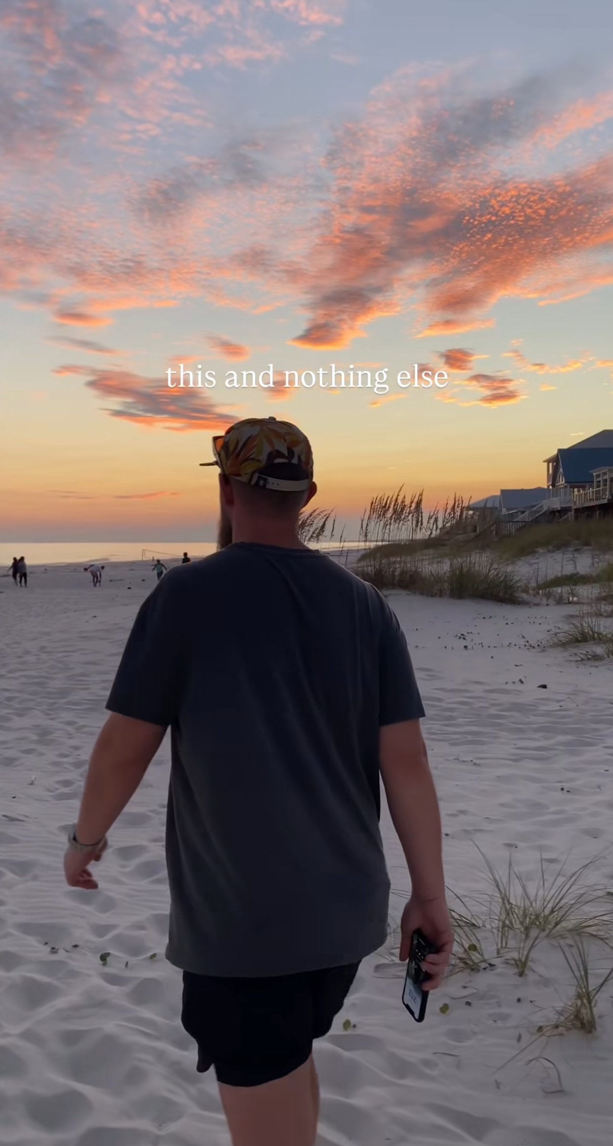 A person walking on a sandy beach at sunset, holding a phone in one hand, with people in the distance, some houses on a dune, and colorful clouds in the sky. Text overlay says 'this and nothing else.'